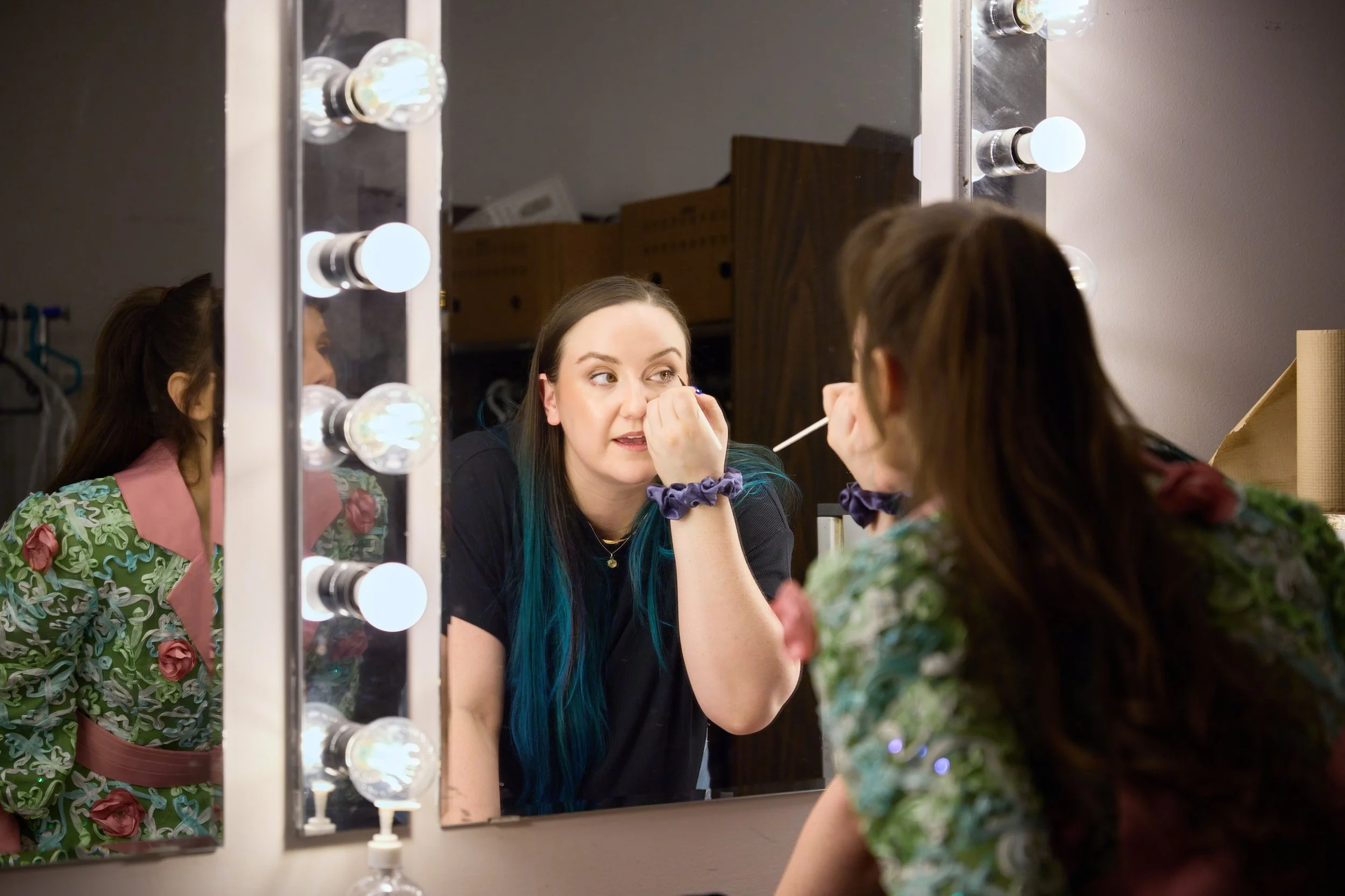 Une jeune femme se maquille devant un miroir avec des ampoules lumineuses, en regardant sa propre reflection.