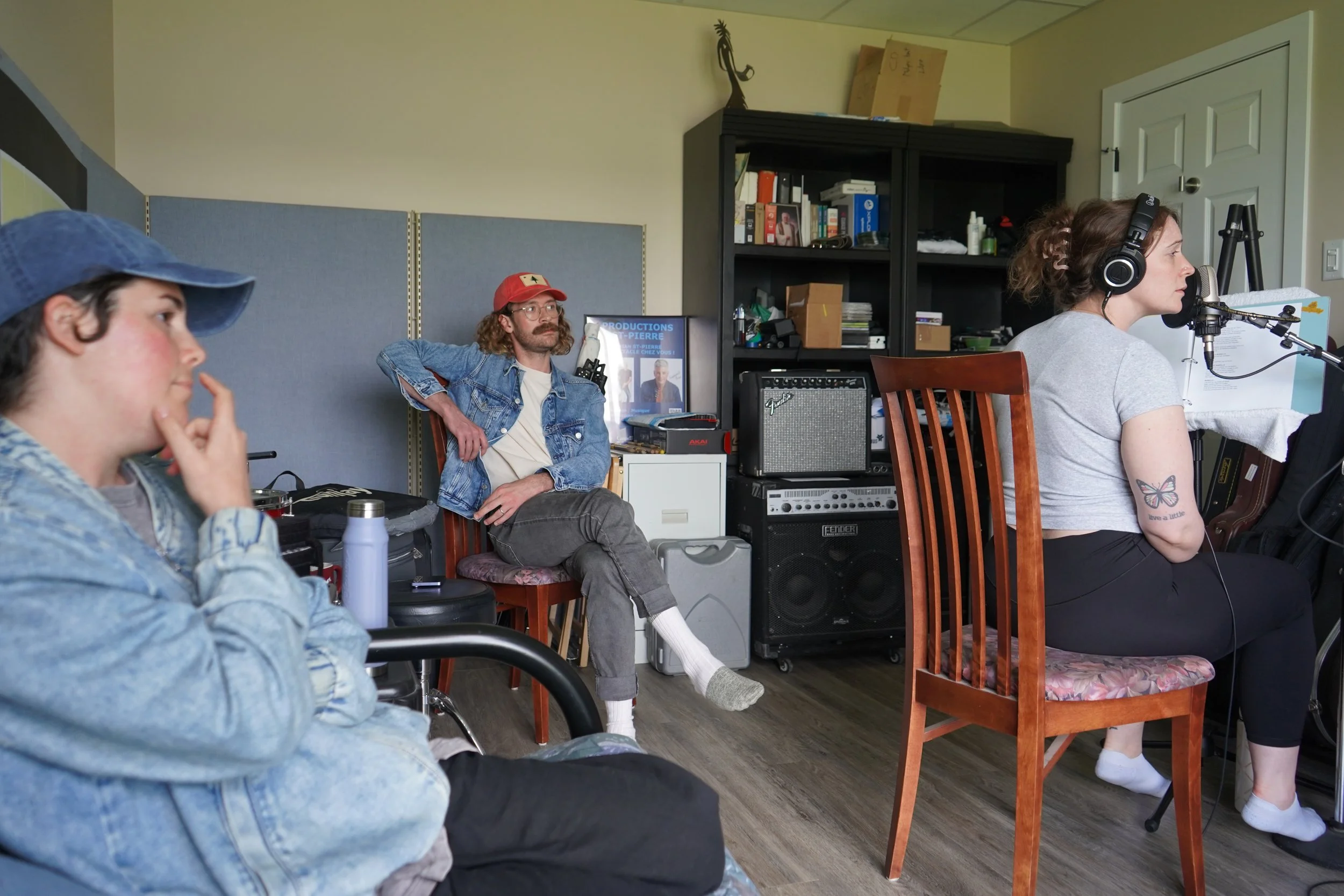 Groupe de personnes dans une pièce, dont une femme utilisant un microphone, une autre personne souriante portant une casquette rouge, et une autre assise en face, avec des étagères remplies de livres et d'équipements audio.