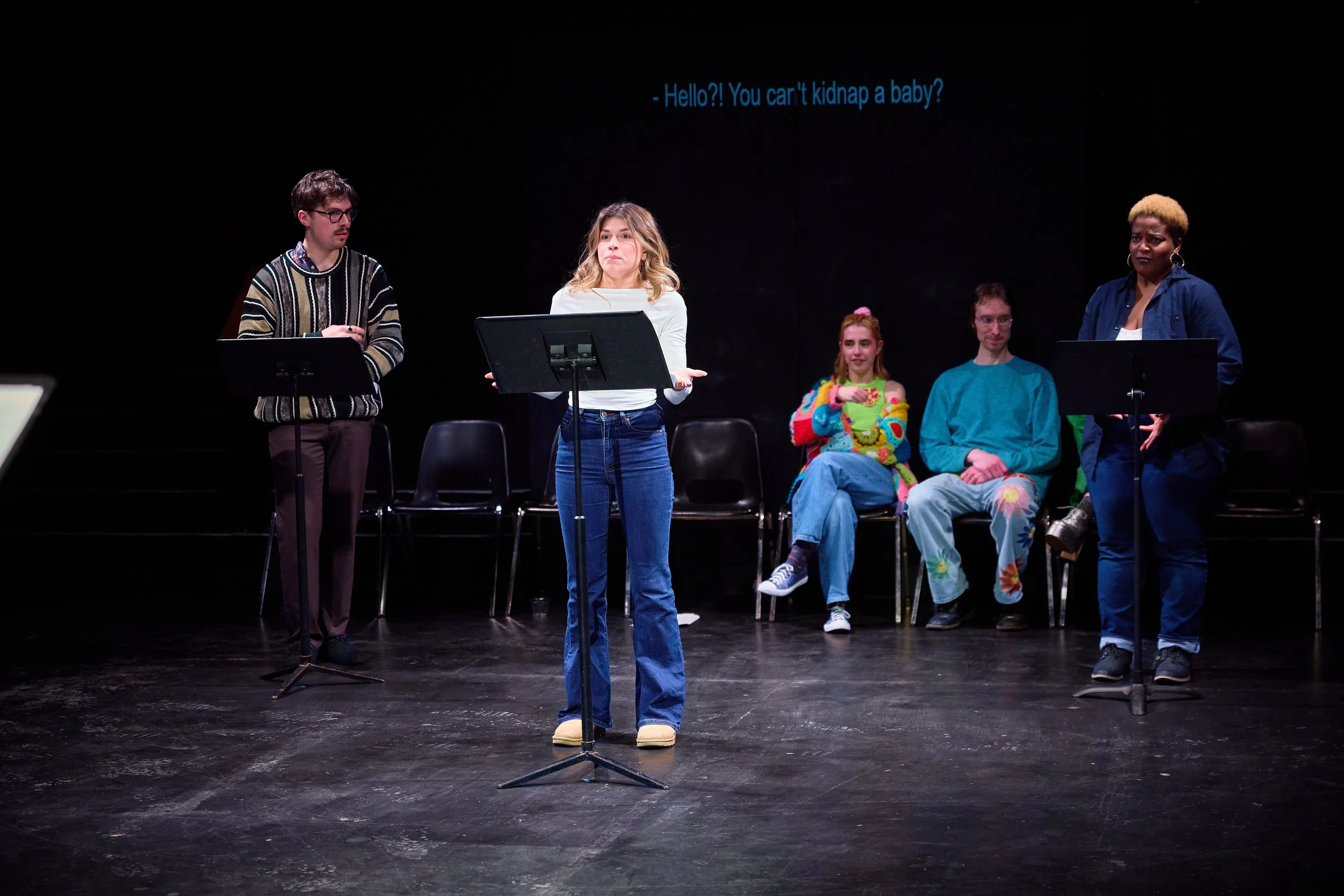 Des personnes se produisent sur scène, avec une femme qui parle au micro, et quatre autres personnes assises derrière elle. Un écran en arrière-plan affiche le texte : 'Hello?! You can't kidnap a baby?'.