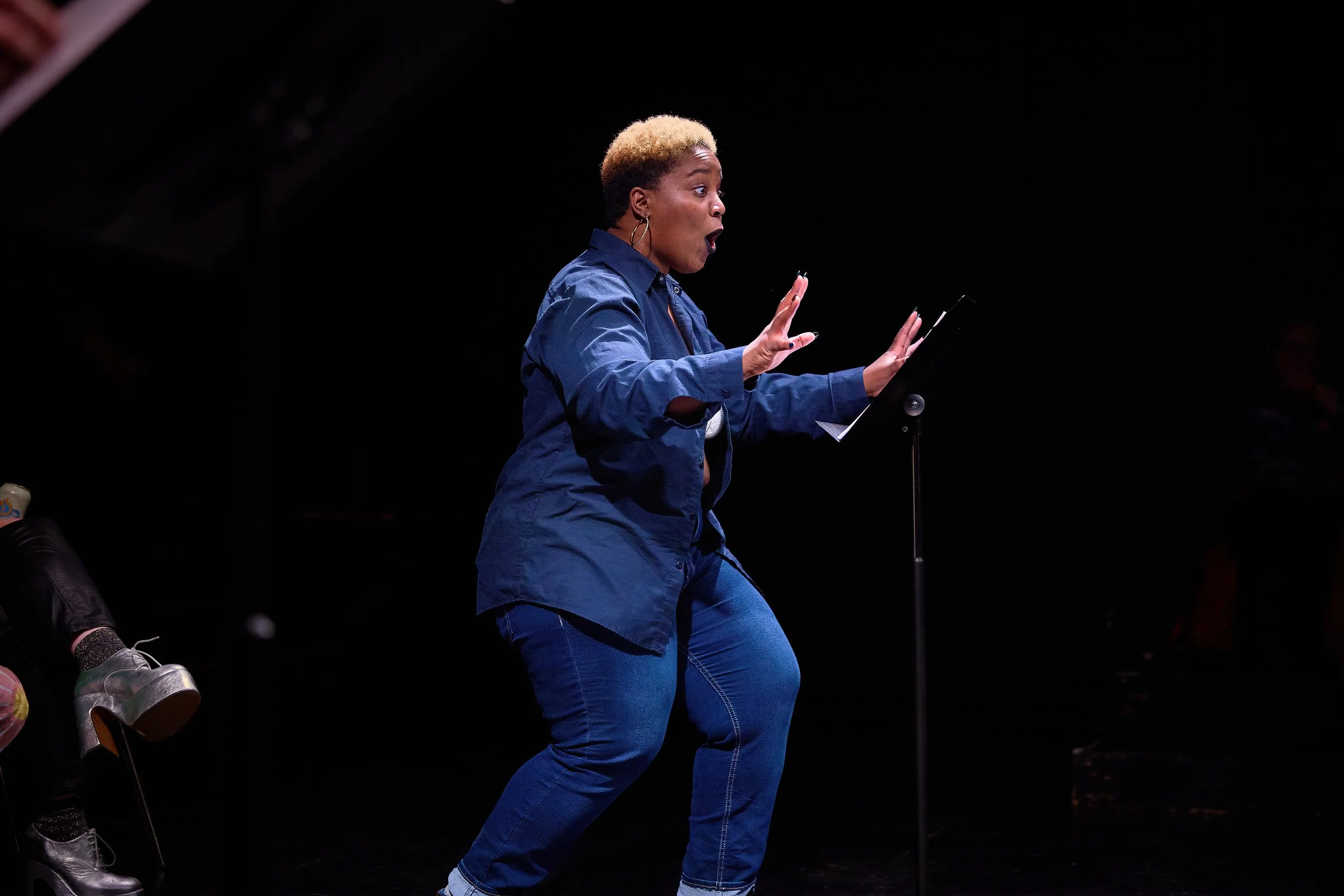 Une femme avec des cheveux blond foncé et une veste bleue, en pleine performance sur scène, gesticulant et semblant s'exprimer avec vivacité.
