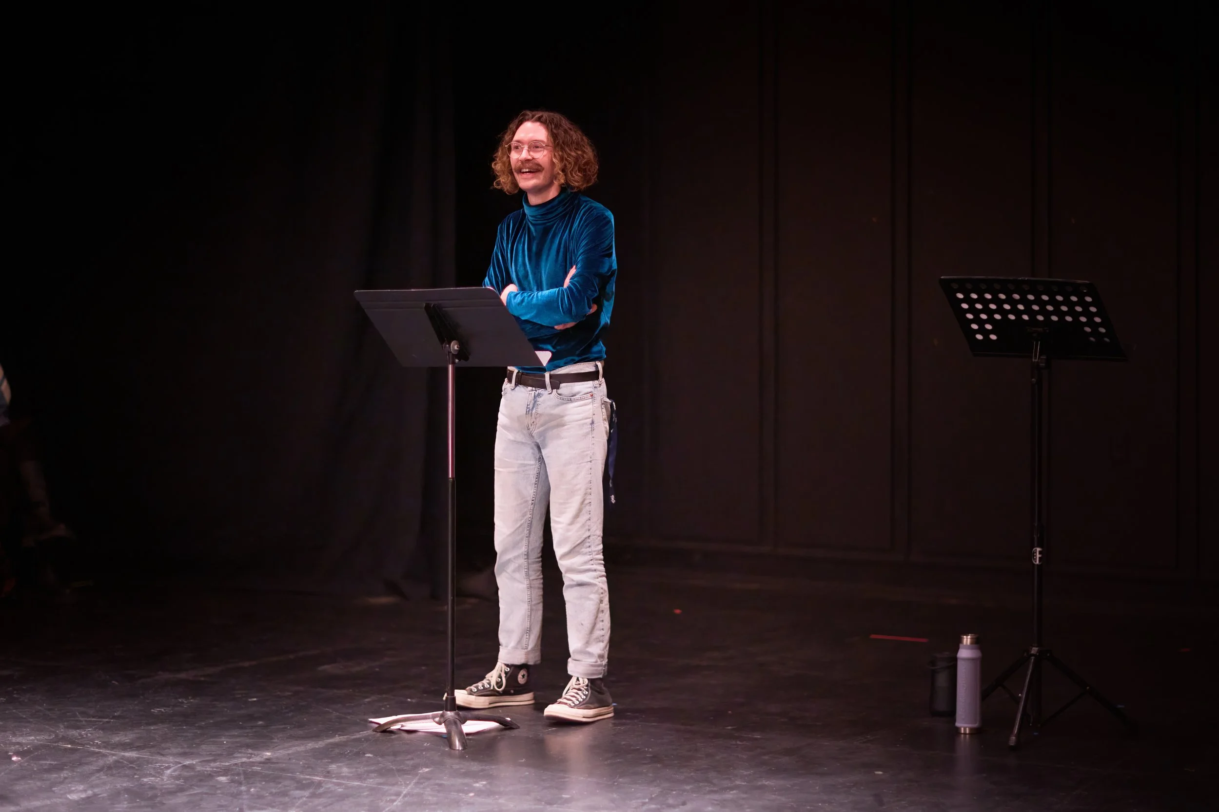 Homme avec cheveux bouclés et moustache, portant un vêtement bleu en velours, se tenant sur scène avec une bâche de lecture, souriant.