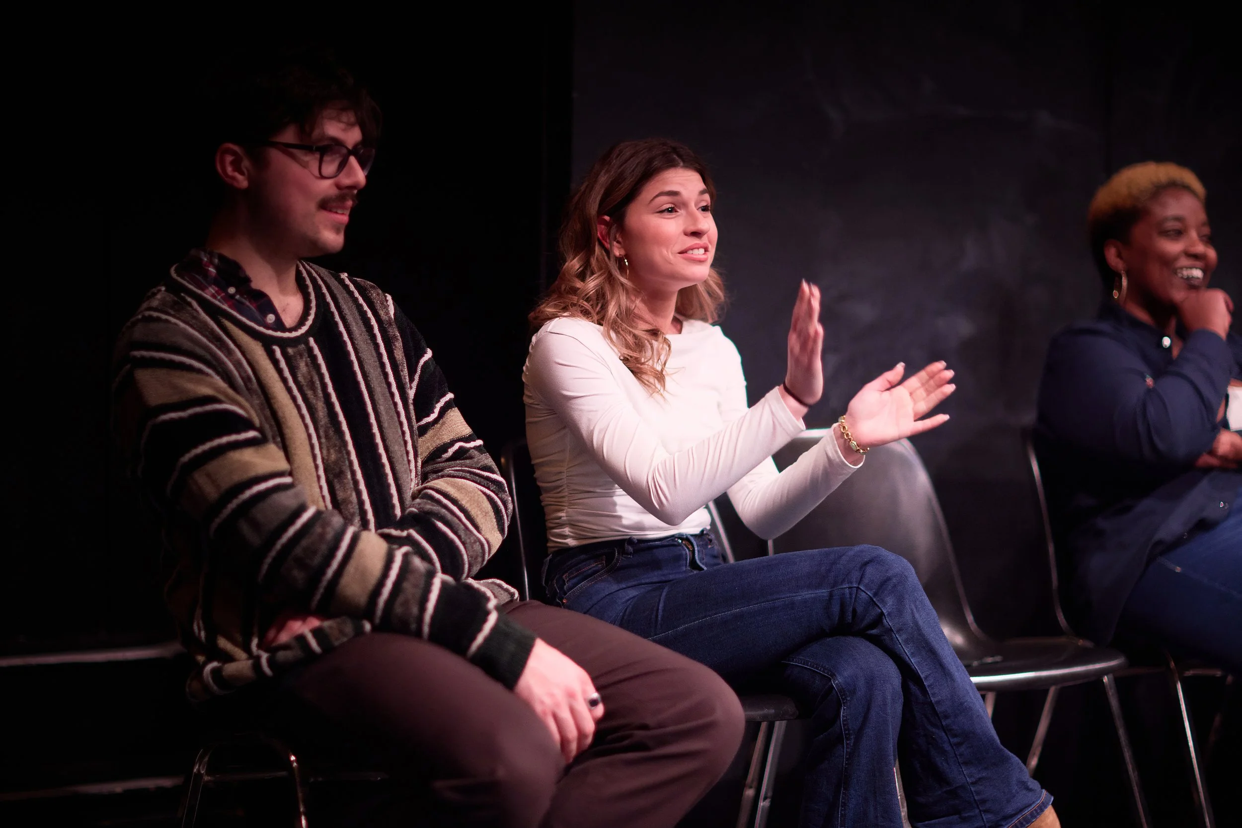 Trois personnes assises en ligne dans un théâtre ou un studio en train de parler ou de réagir, avec un mur noir en arrière-plan.