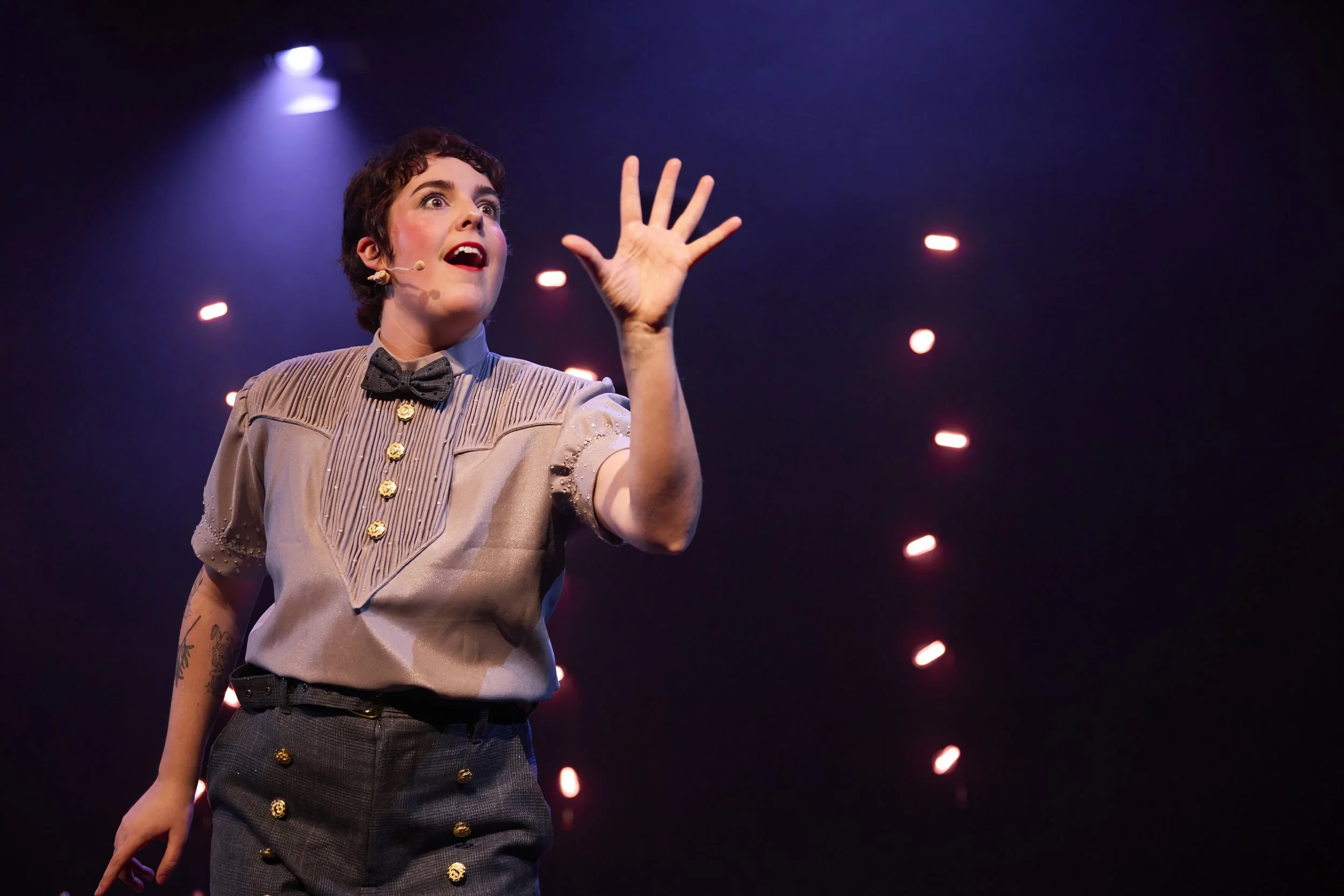 Créations In Vivo. Une femme sur scène, gesticulant avec une expression enthousiaste, sous un éclairage violet et rose, portant une tenue vintage avec des détails en or.