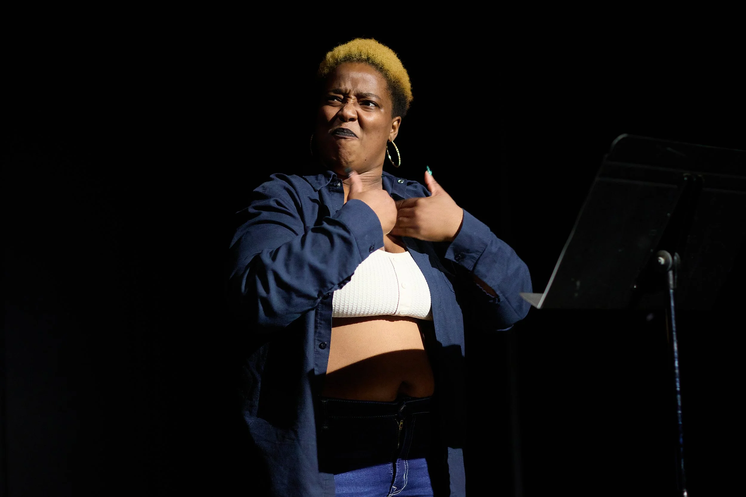 Une femme avec des cheveux blonds courts, portant une chemise bleue, une brassière blanche et un pantalon noir, gesticulant avec une expression faciale intense, sur scène avec un pupitre ou une partition à côté.