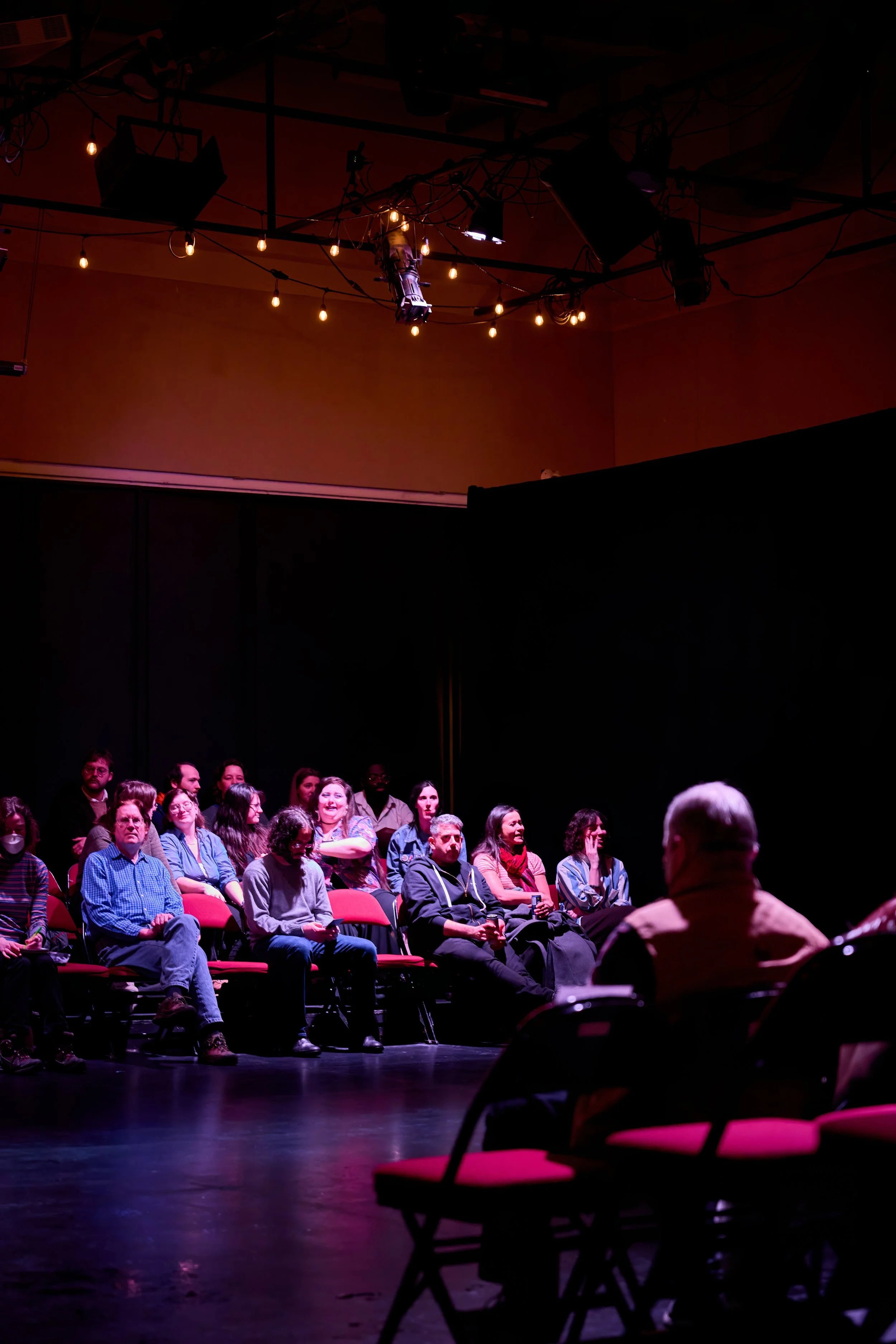 Public assis dans une salle sombre, probablement un théâtre ou une salle de spectacle, regardant un performeur ou un speaker sur scène, avec une lumière tamisée et une ambiance intimiste.