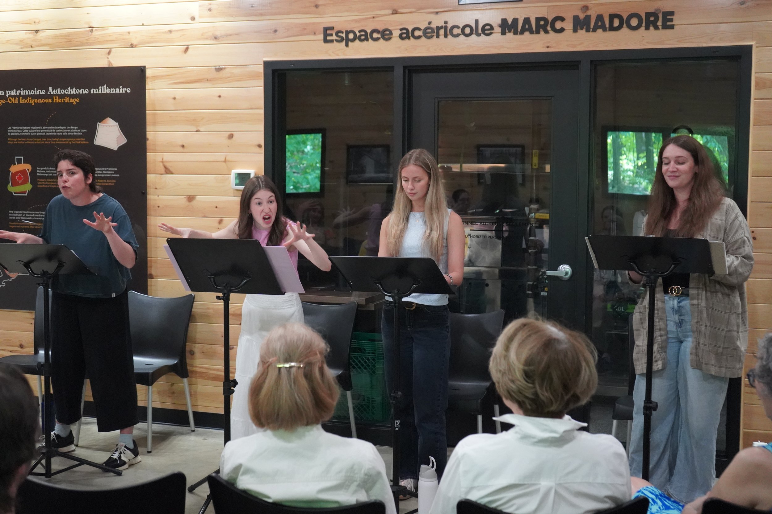 Quatre femmes chantant ou parlant devant un public, dans une pièce avec murs en bois, quelques partitions sur des pupitres, et un panneau informatif sur la gauche. L'inscription indique 'Espace acéricole MARC MADORE'.