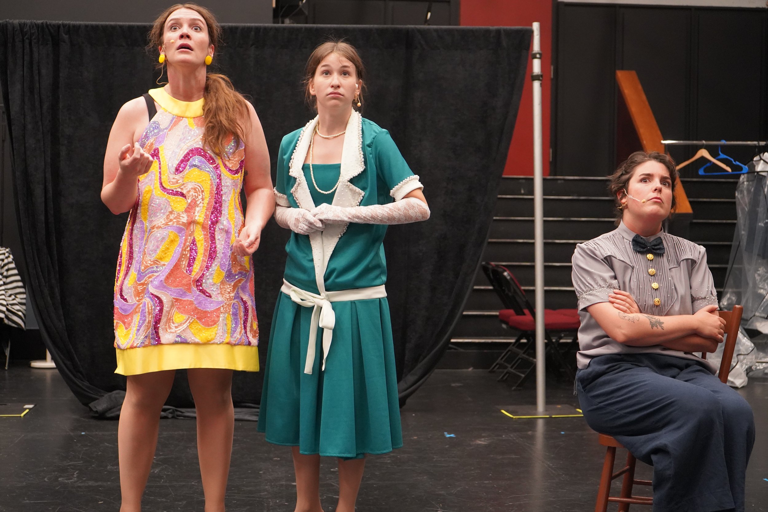 Trois femmes en costumes vintage sur scène, dont deux en première ligne et une assise à droite, dans un théâtre ou une salle de spectacle.