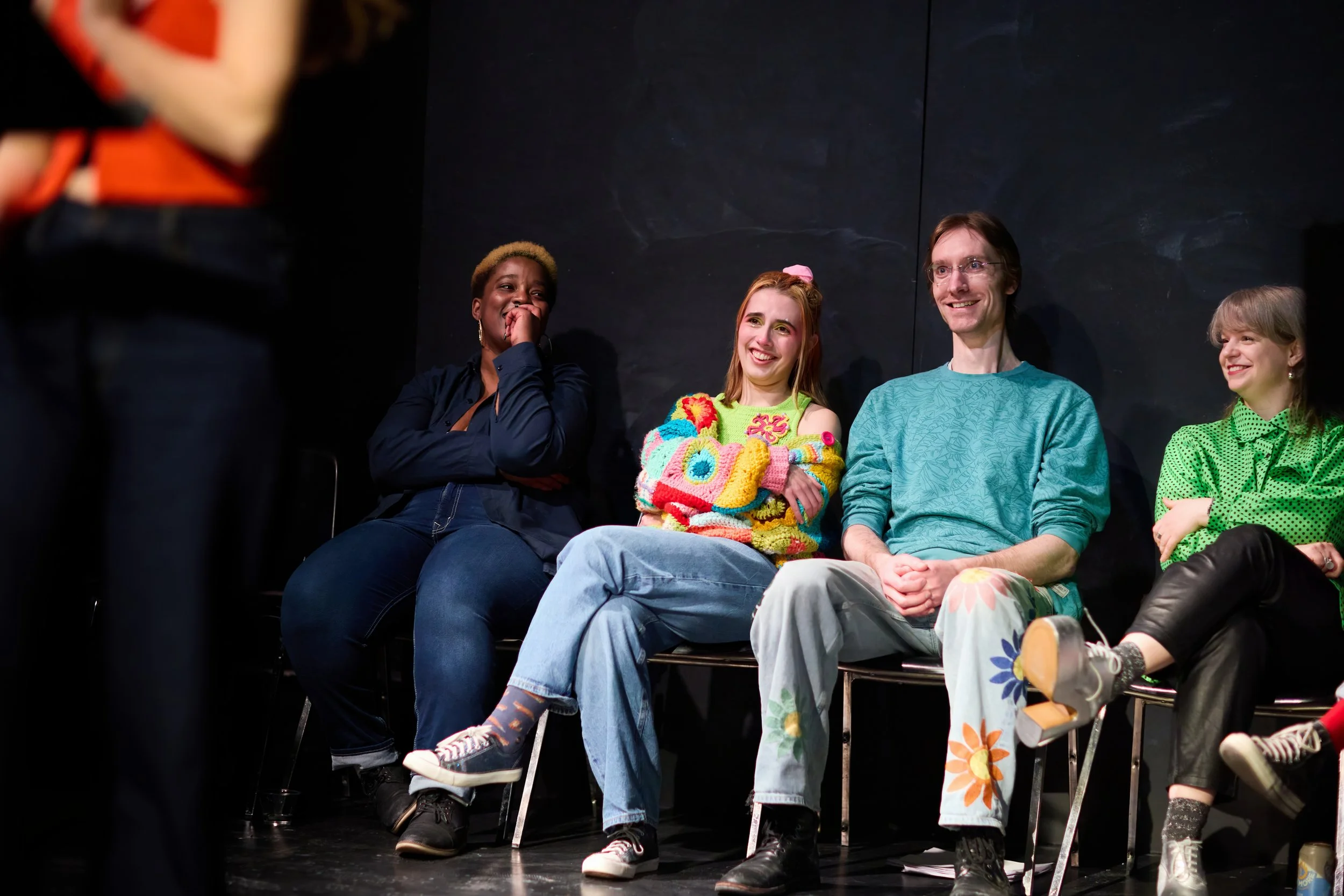 Groupe de personnes assises en mode décontracté, souriantes, dans une pièce sombre, probablement en train de discuter ou d'assister à un événement humoristique ou artistique.