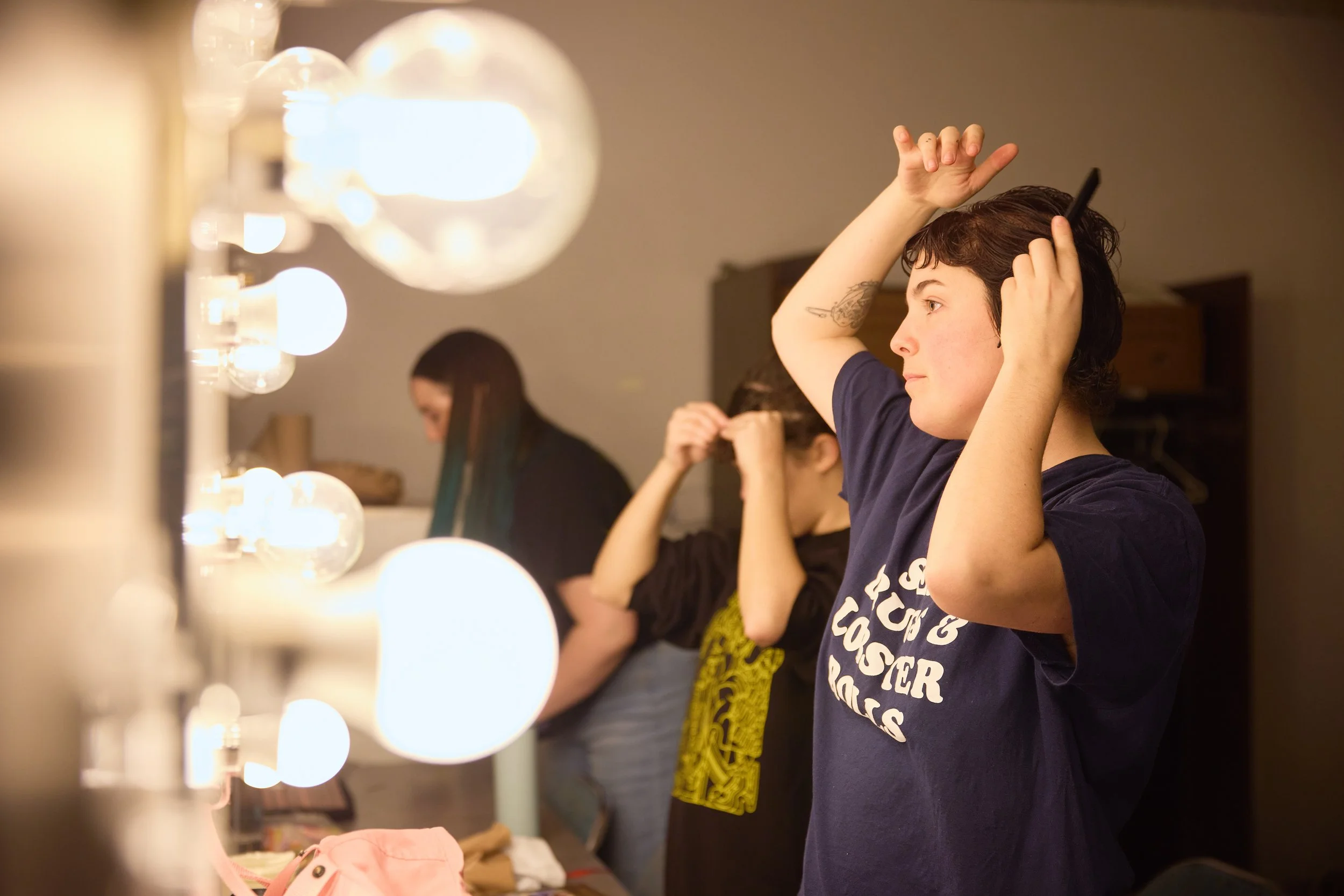 Une jeune femme avec des cheveux courts et une T-shirt bleu est en train de se coiffer en regardant dans un miroir, avec d'autres personnes dans le fond. Elle se tient devant un éclairage de maquillage avec des ampoules.