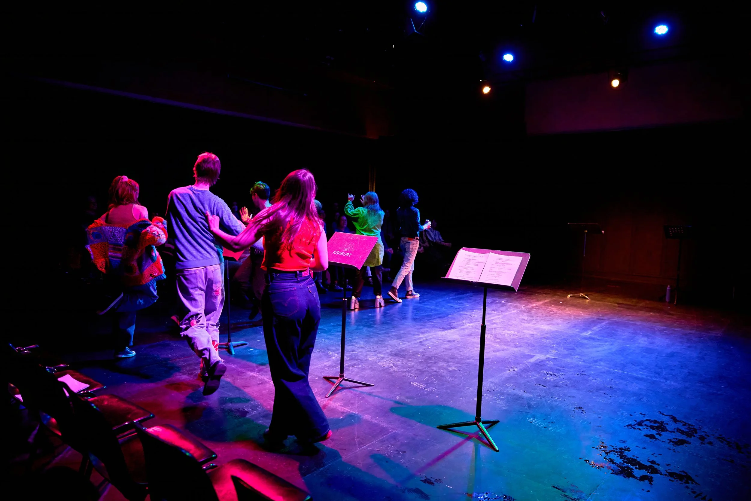 Groupe de personnes en train de danser sur scène sous des lumières colorées, avec des partitions de musique sur des pupitres
