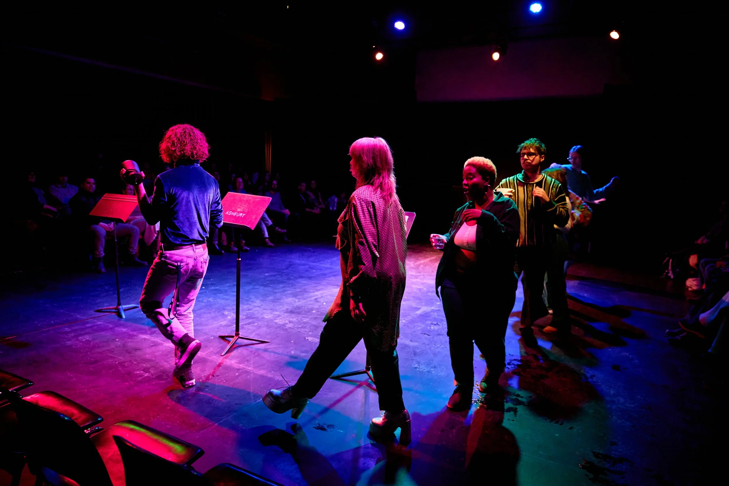 Groupe de personnes en spectacle sur scène avec éclairages colorés, public assis en arrière-plan