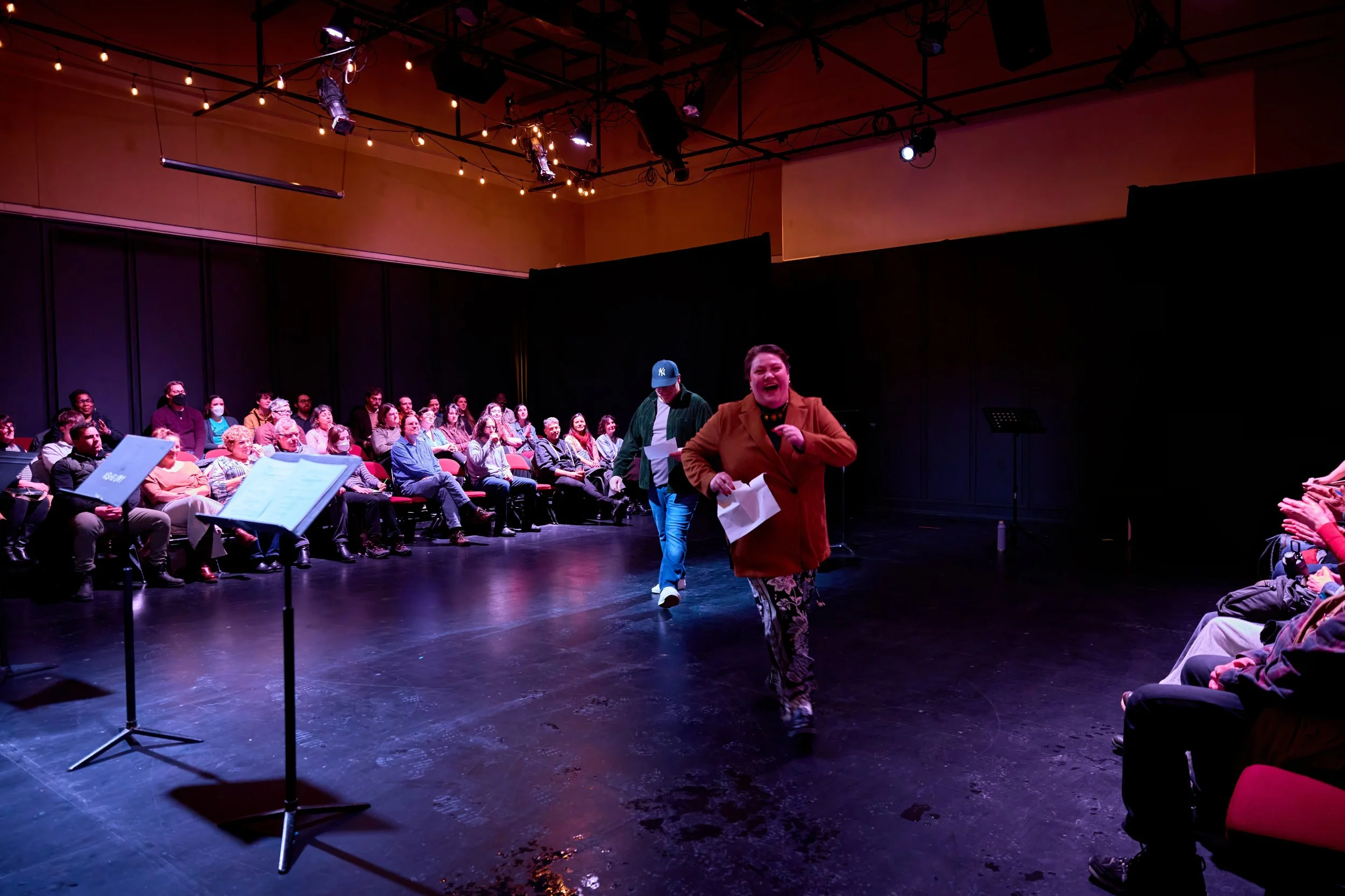 Une scène de théâtre ou de spectacle avec un public assis en face d'une scène sombre. Une femme souriante et en train de parler ou de chanter se trouve au centre, entourée d'autres personnes, dans une ambiance éclairée chaleureusement.