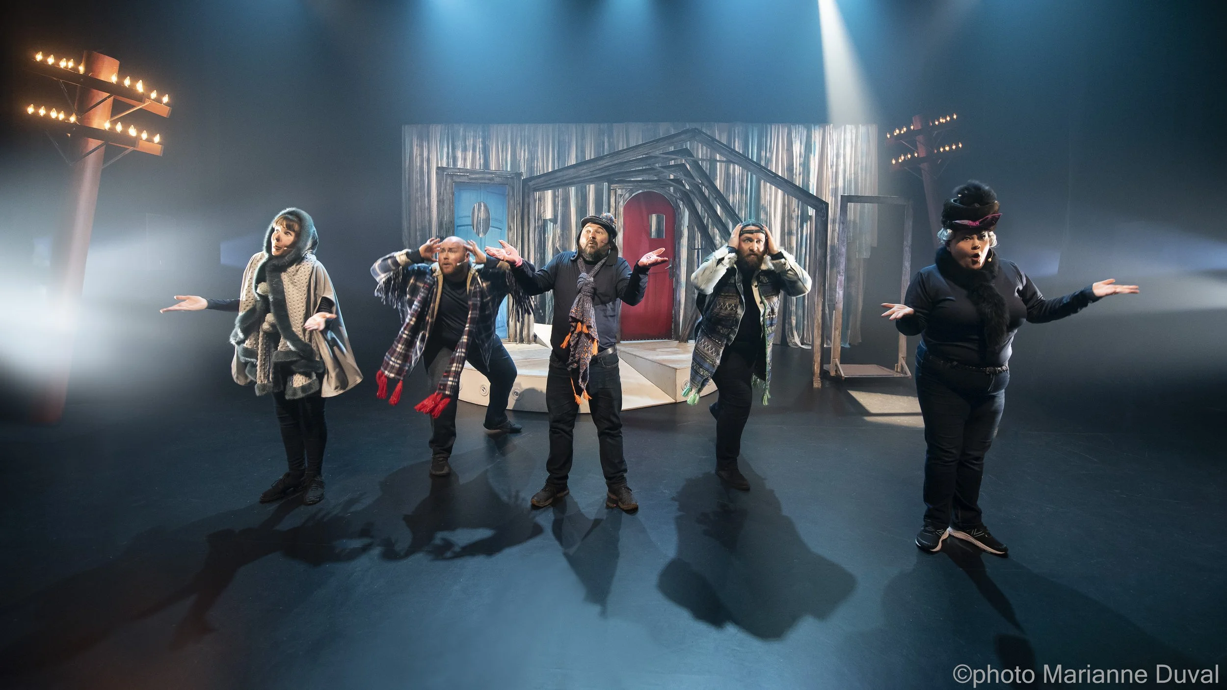 Groupe de cinq acteurs en costume sur scène, en train de jouer une scène de théâtre, avec fond en bois, portes rouges, et lumières de scène.