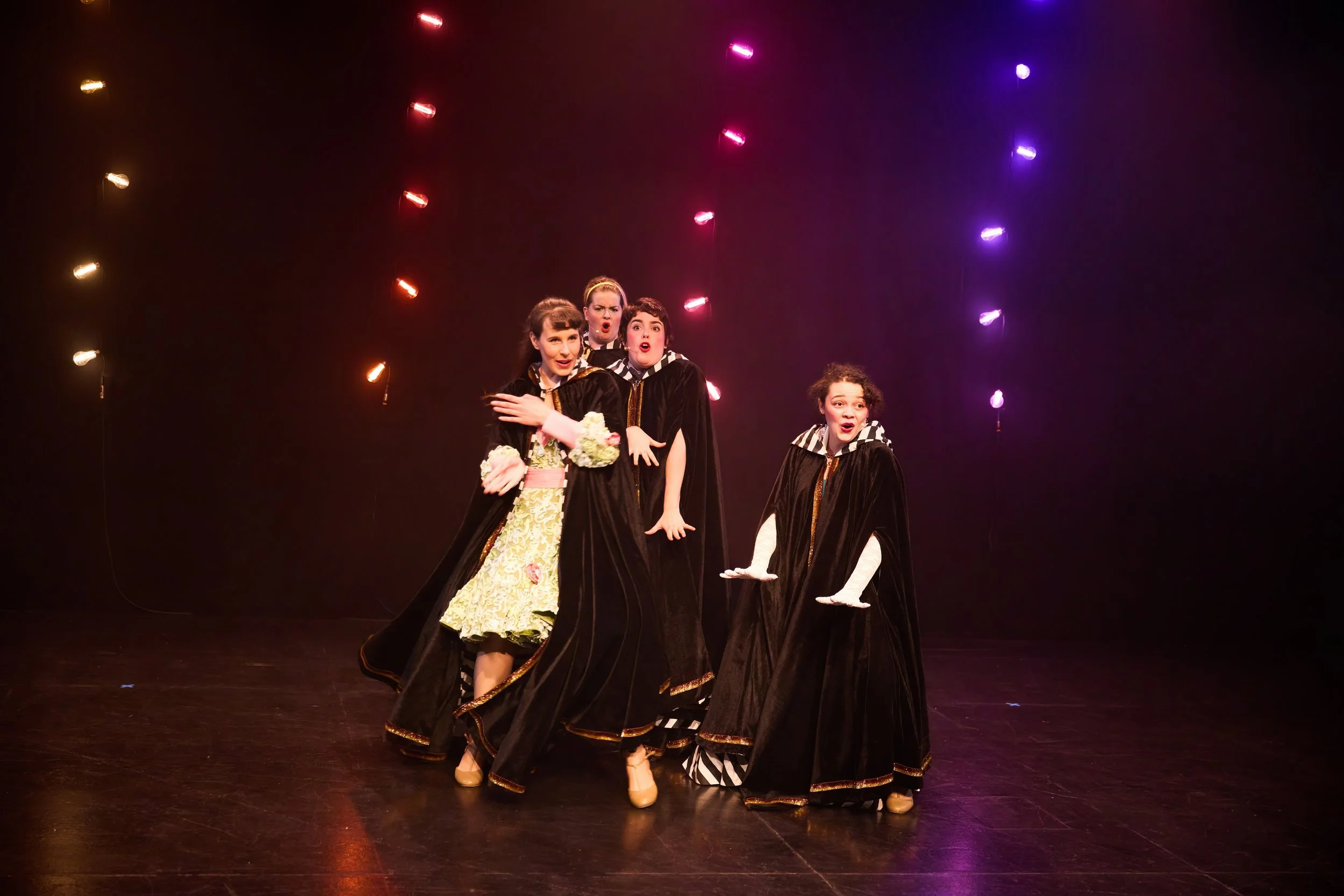 Quatre femmes en costumes de théâtre noir, en pleine scène, sous des lumières colorées, exprimant des émotions variées.