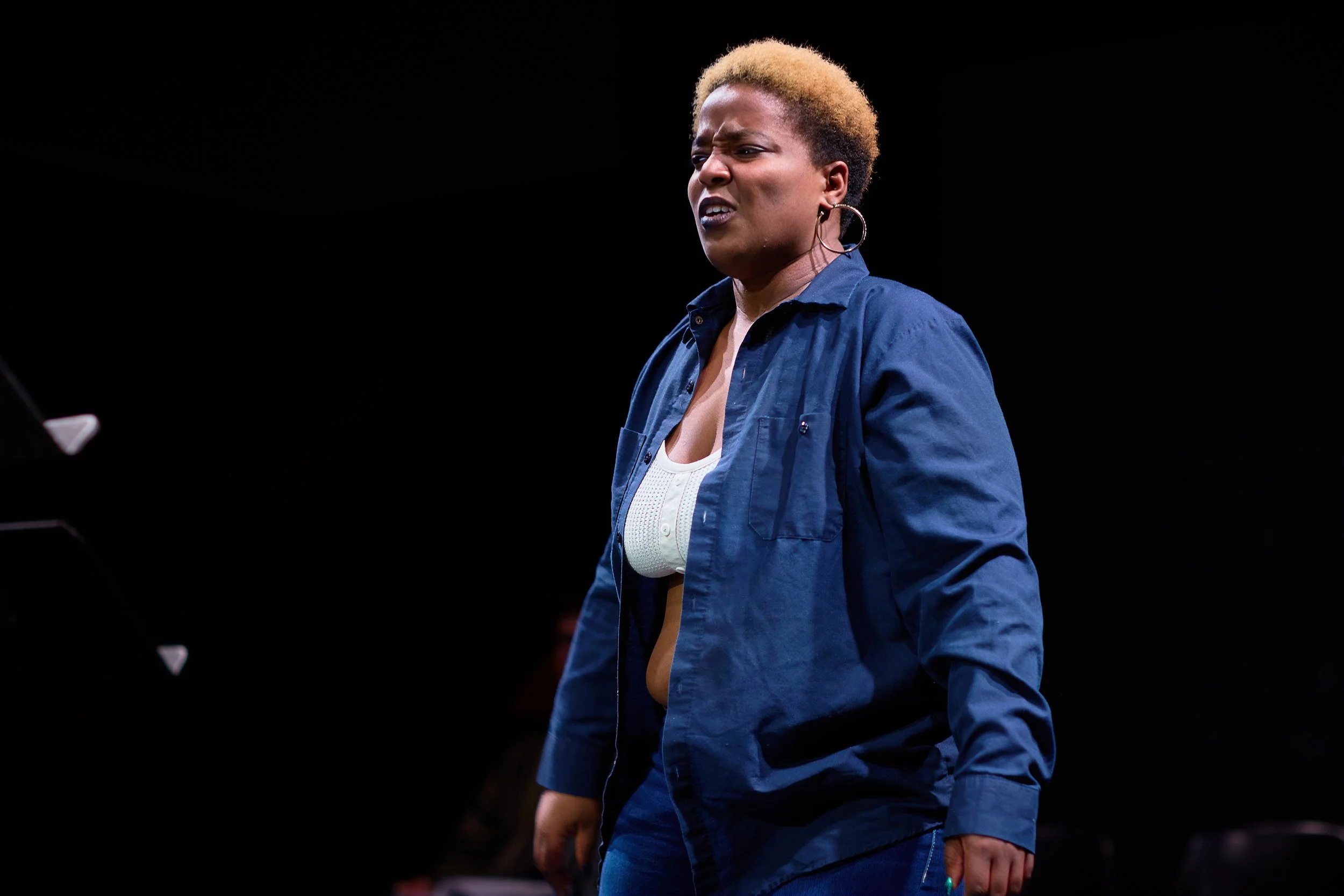 Une femme avec des cheveux courts blond foncé, portant une veste bleue et une blouse blanche, a une expression intense sur le visage, probablement en train de parler ou de performer sur scène