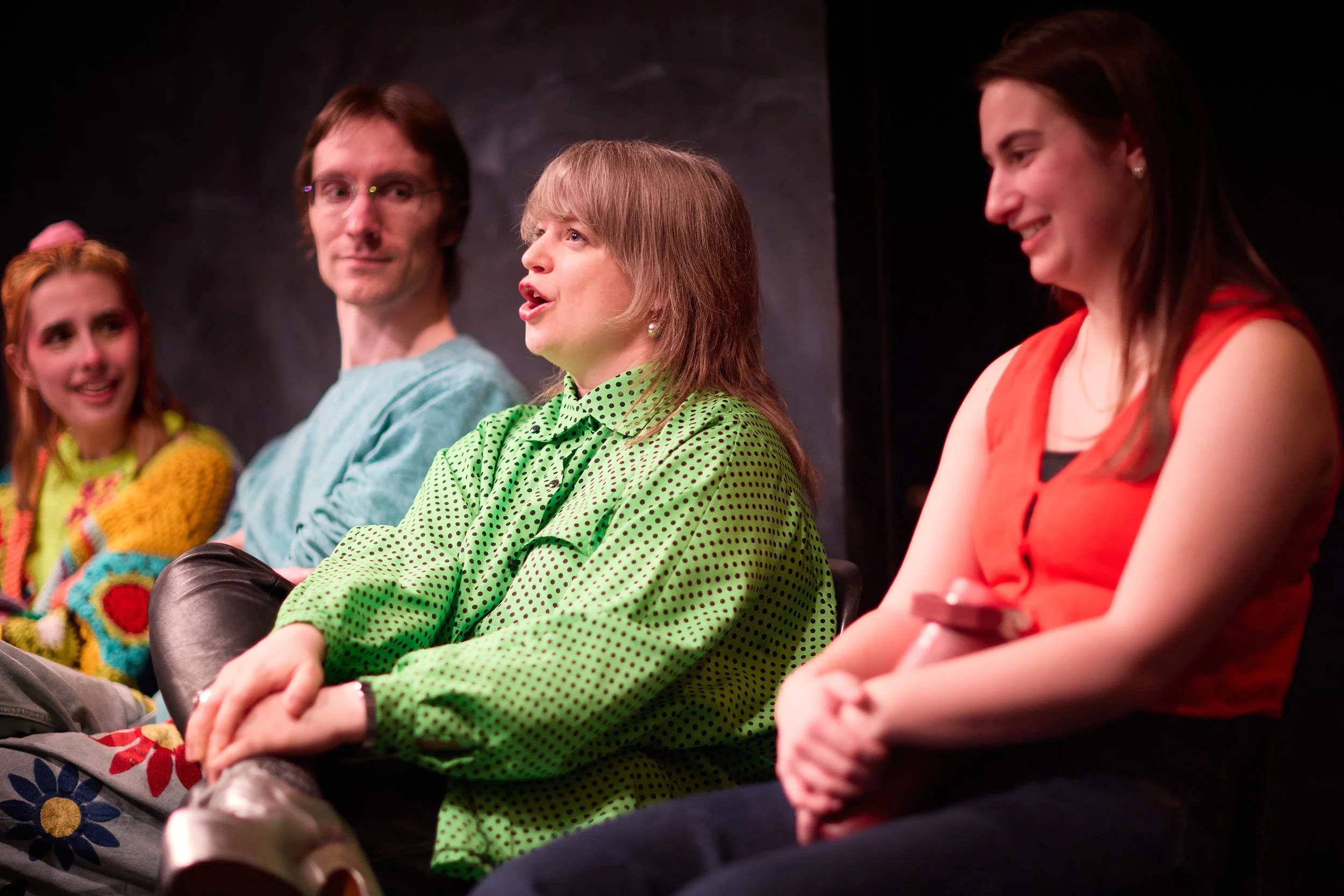 Groupe de quatre personnes assises sur scène lors d'une pièce de théâtre, discutant ou écoutant, avec un fond sombre.