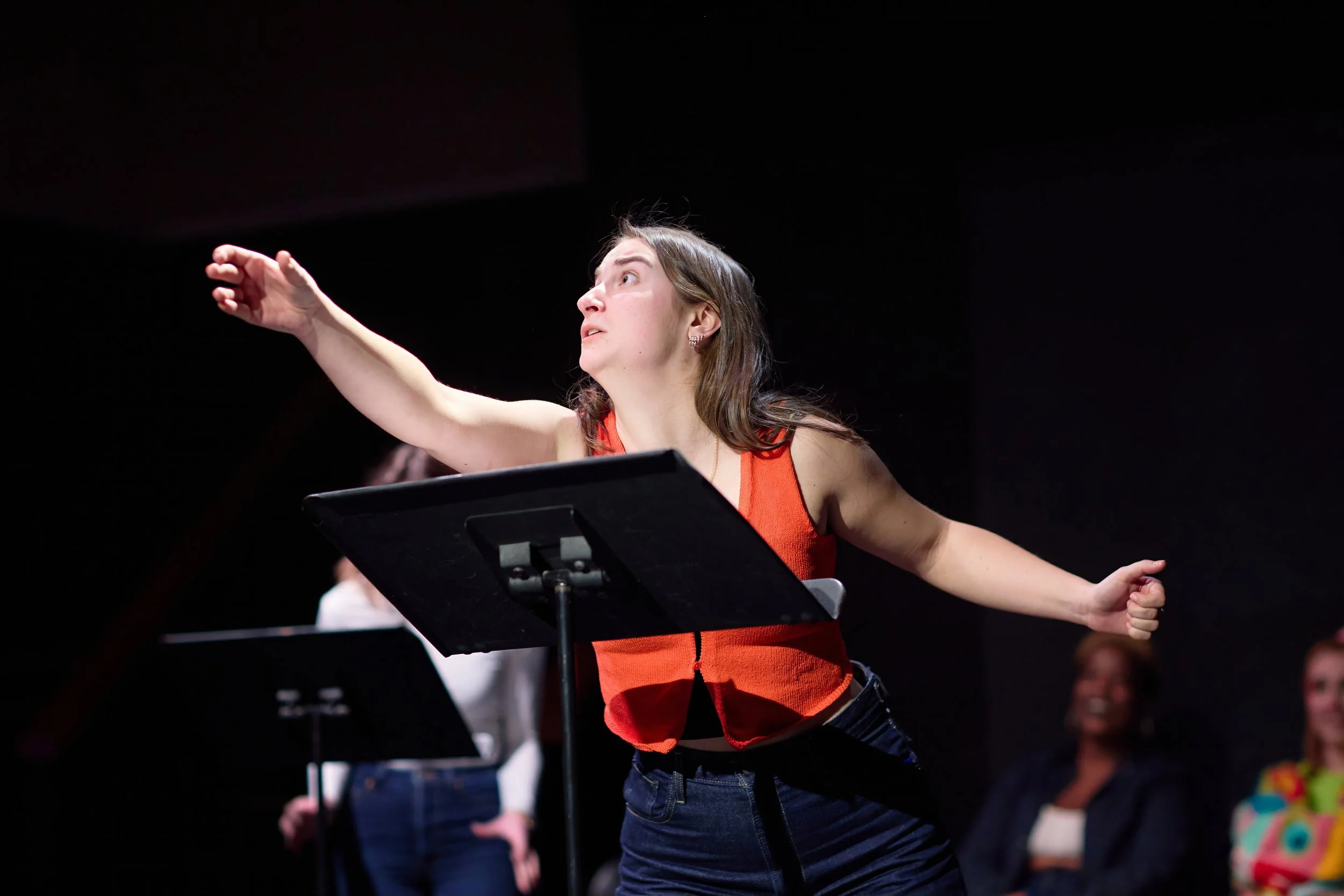 Une jeune femme dirigée une répétition ou un spectacle, gesticulant avec enthousiasme derrière un pupitre, avec d'autres personnes en arrière-plan.