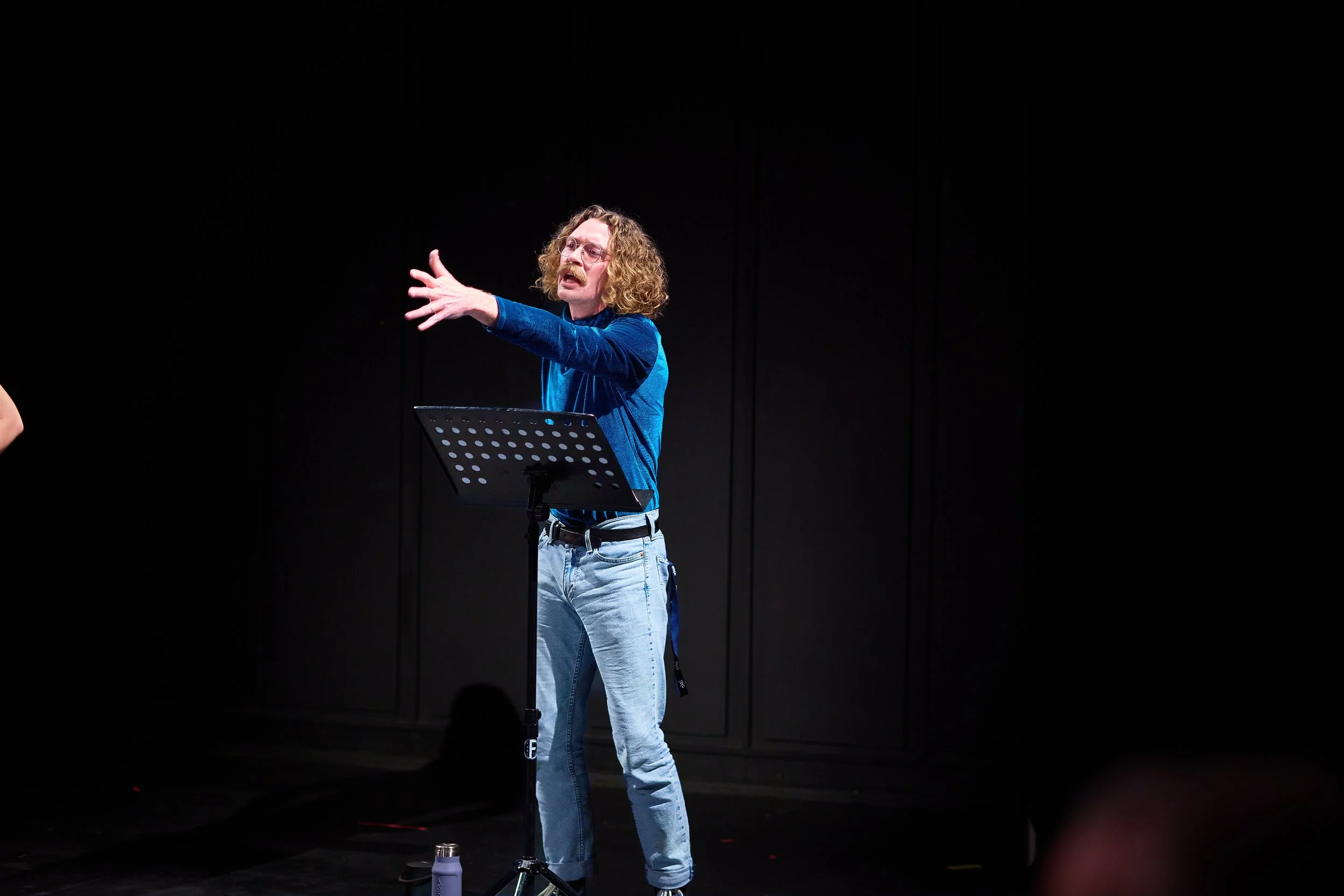 Un homme avec des cheveux bouclés et une moustache, vêtu d'un pull bleu et de jeans, parle ou chante avec enthousiasme sur scène, avec un pupitre ou un support de musique devant lui, dans un environnement sombre.