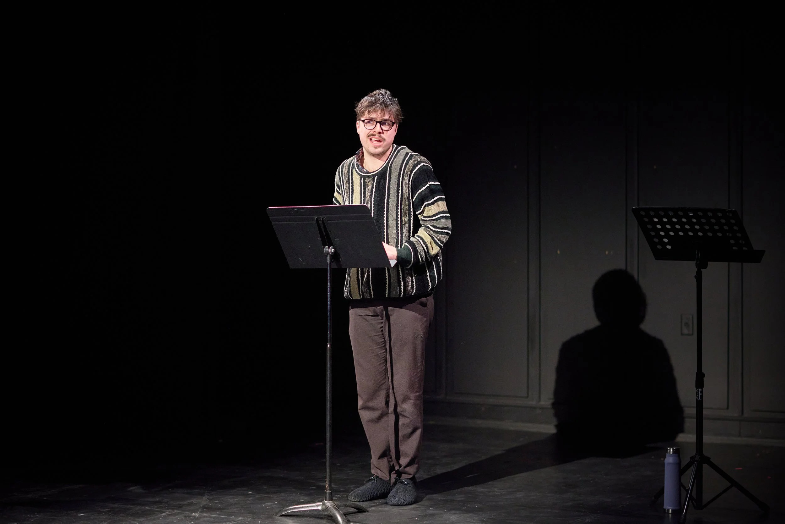Un homme parle sur scène avec un pupitre, en arrière-plan un espace sombre, un support à partition et un reflet de l'homme, se tenant dans une pièce sombre et vide.