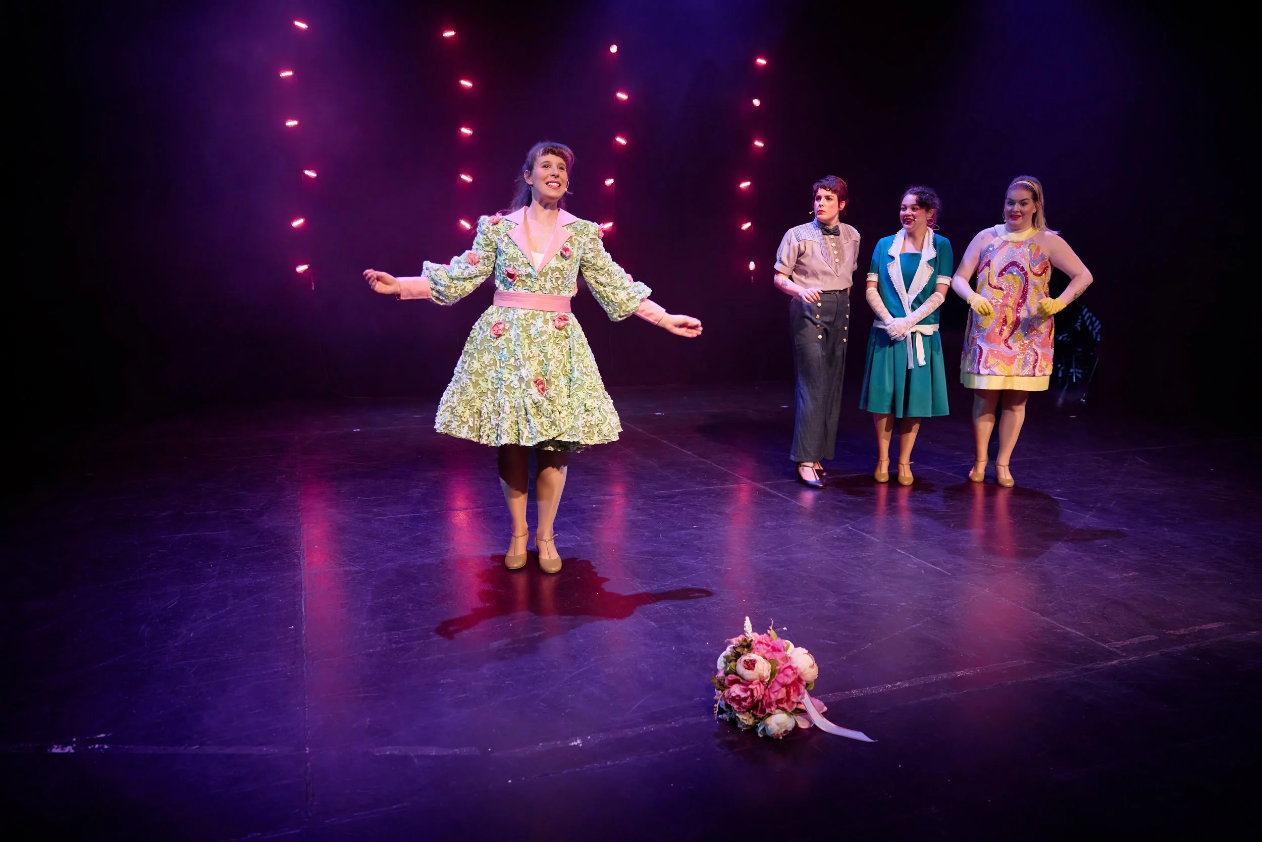 Scène de théâtre avec quatre acteurs en costumes vintage, une femme au centre souriante avec un bouquet de fleurs devant elle, sous un éclairage rose et violet.