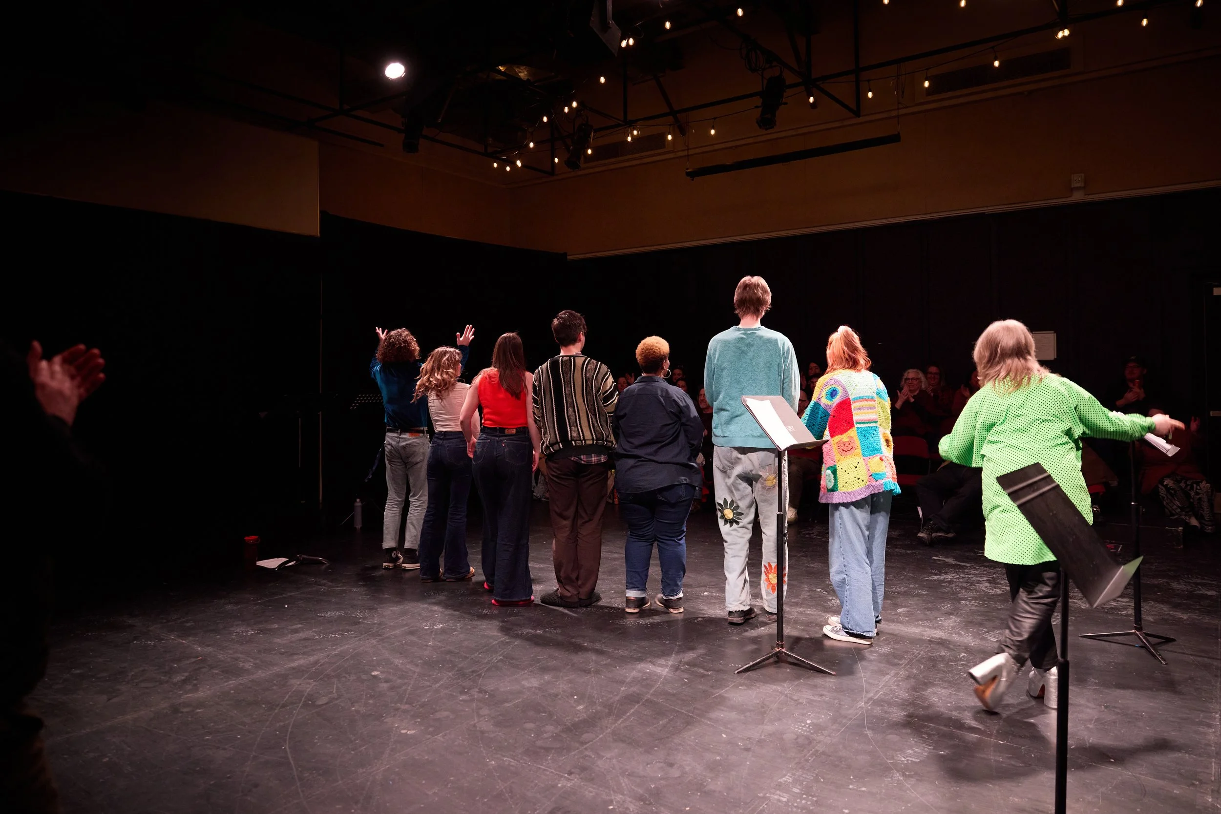 Groupe de personnes sur scène, vue de dos, en pleine performance ou répétition, avec un public assis en arrière-plan, sous des lumières suspendues.