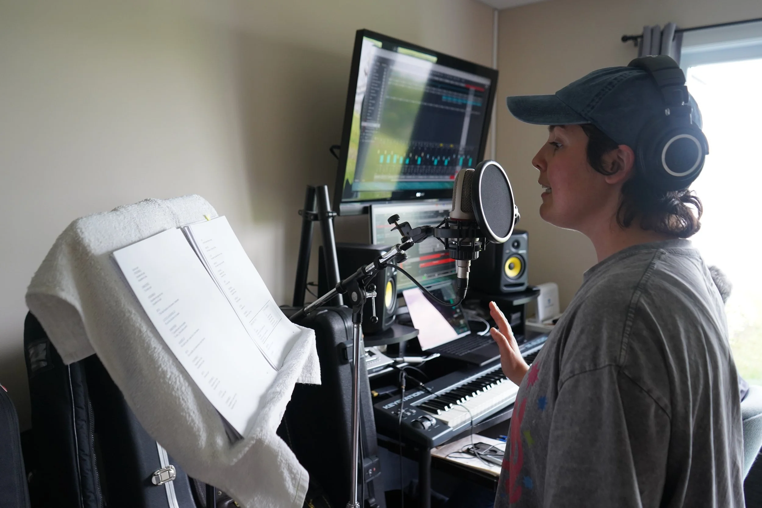 Une femme en train de chanter dans un studio d'enregistrement, portant un casque audio, avec un microphone, un ordinateur, une partition de musique sur un support et un clavier électrique dans la pièce.