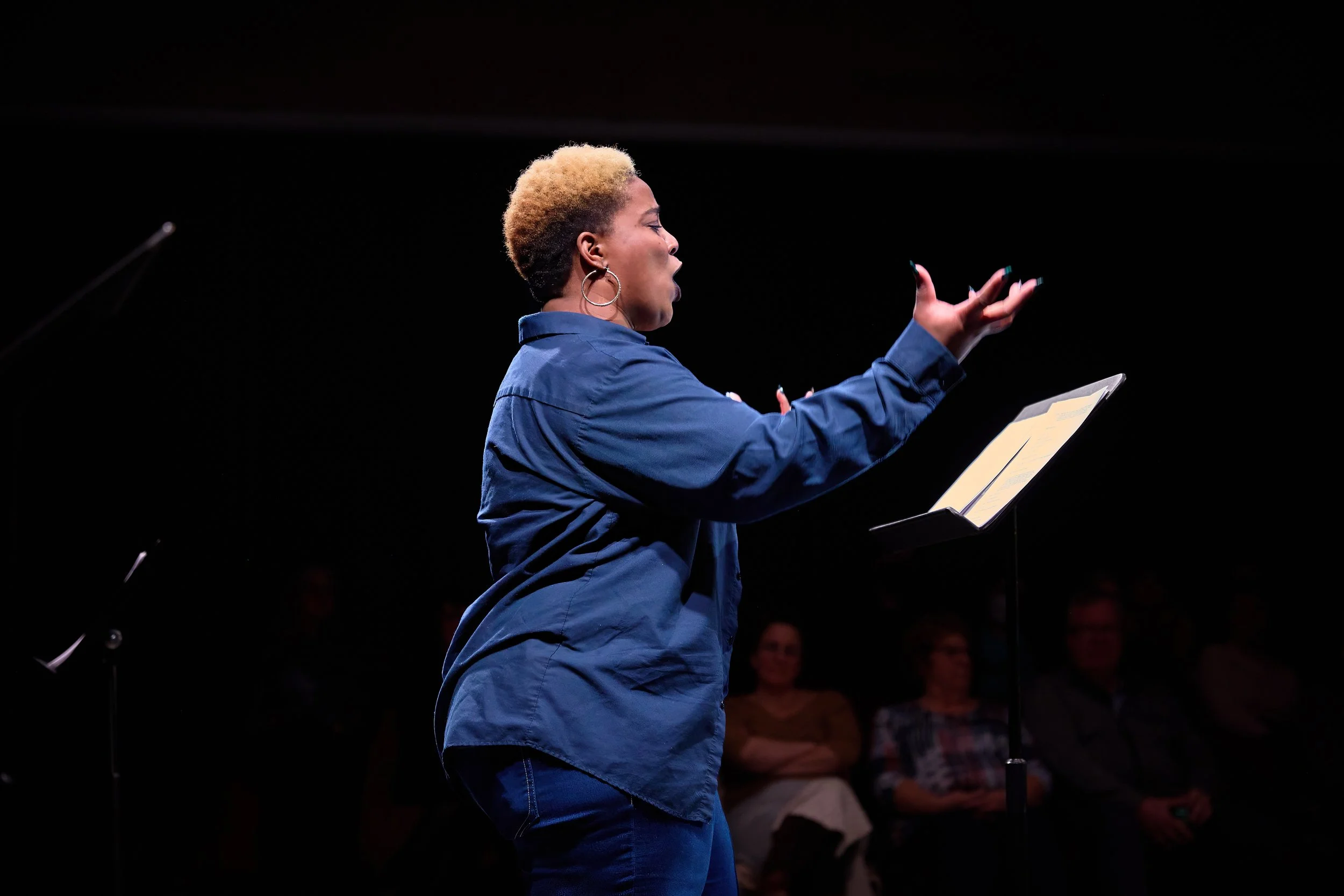 Une femme en chemise bleue parle lors d'une performance devant un micro, avec des spectateurs en arrière-plan.