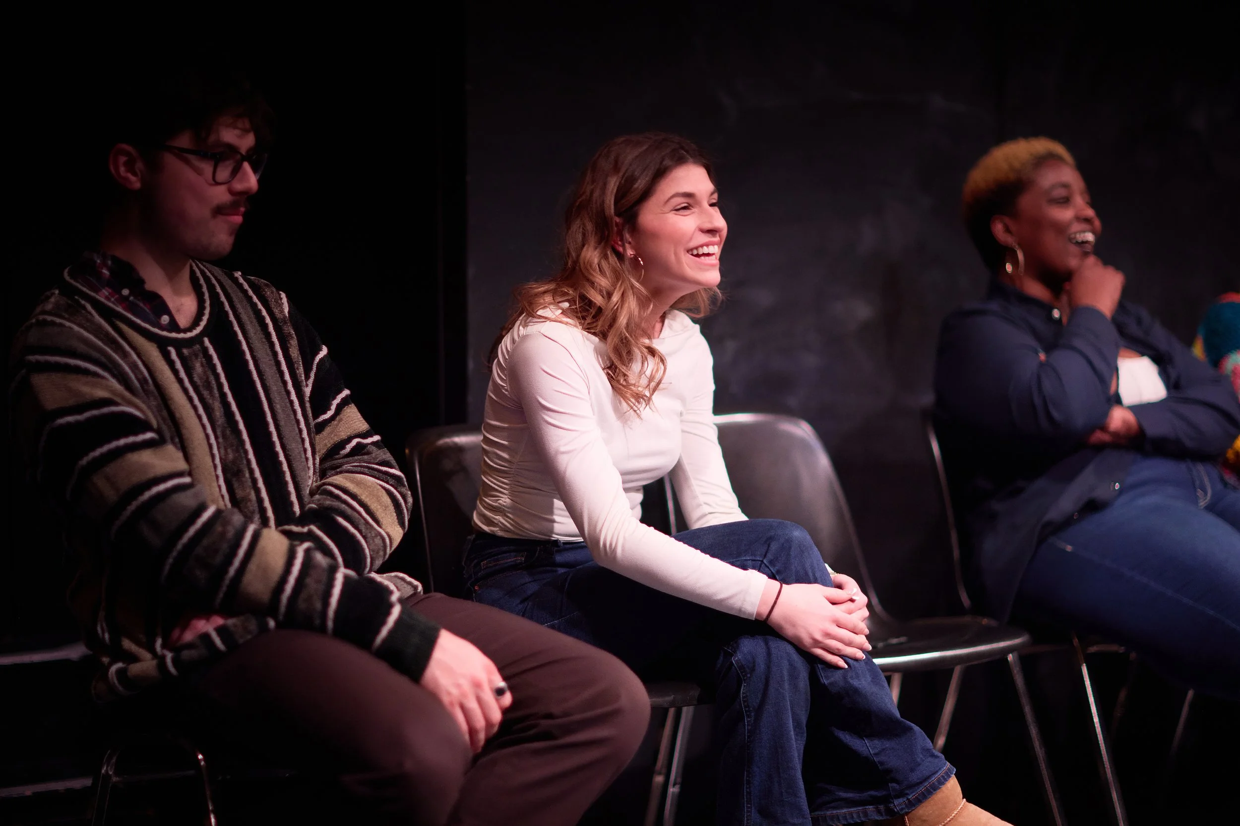 Trois personnes assises sur des chaises dans une pièce sombre, rient et semblent apprécier un spectacle ou une présentation.