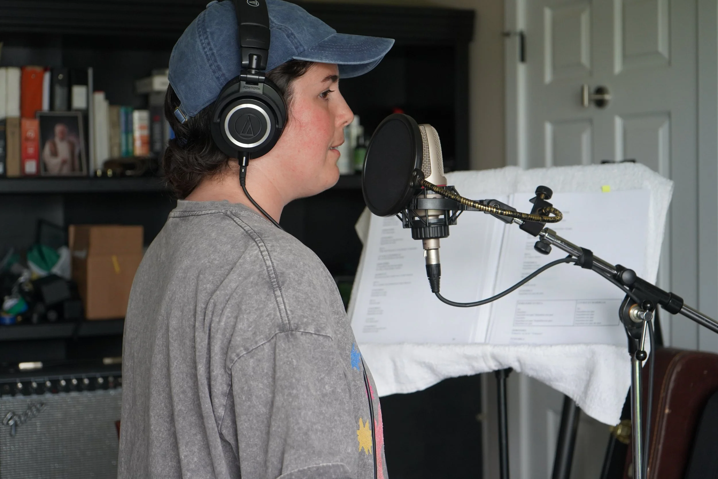 Jeune homme avec casque d’écoute studio enregistrement chante dans un micro de studio, arrière-plan avec étagère et partitions musicales.