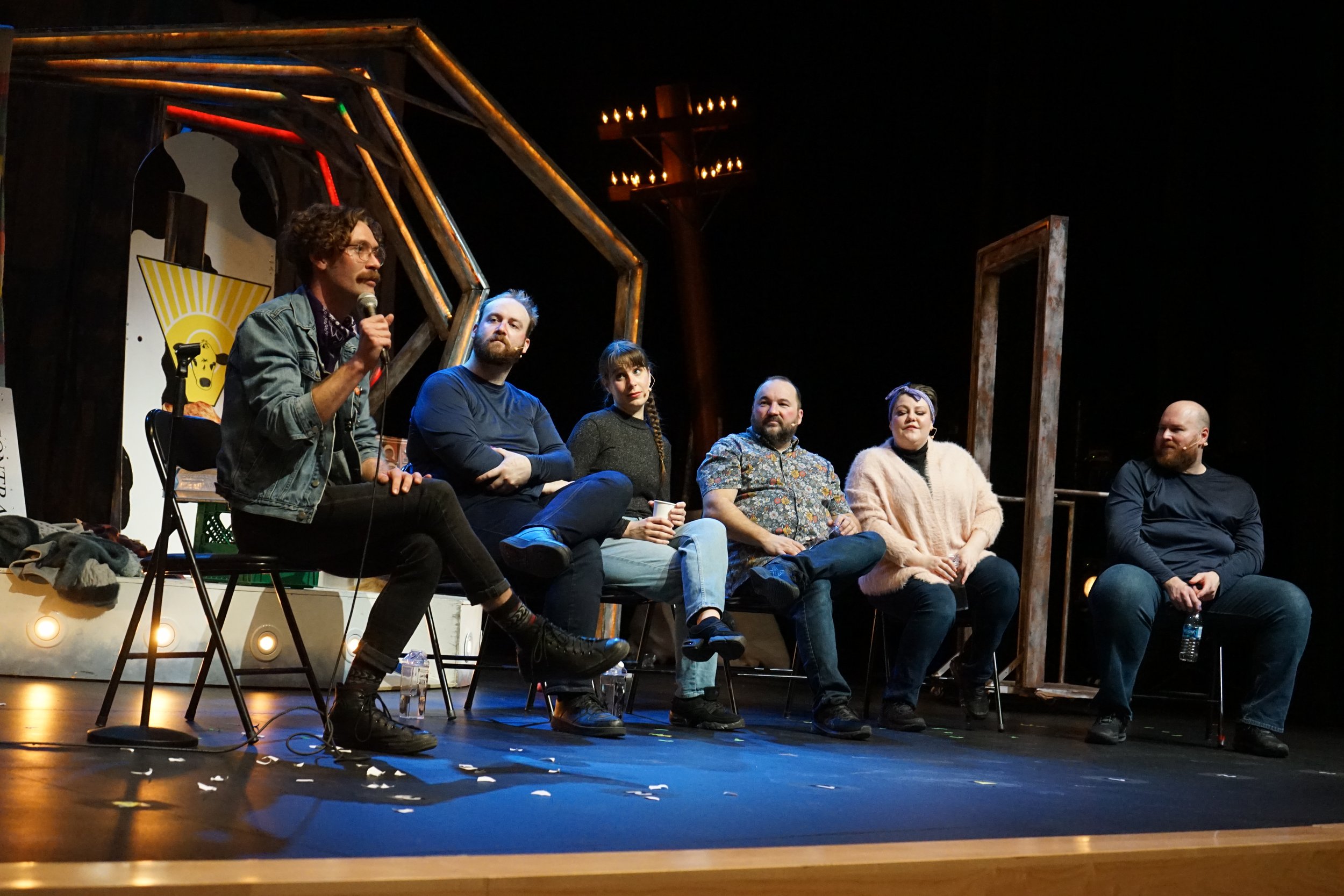Un groupe de six personnes assises sur scène lors d'une discussion. La personne la plus à gauche parle en tenant un micro. La scène est décorée avec une structure métallique et un panneau avec un visage, éclairée par des lumières suspendues.