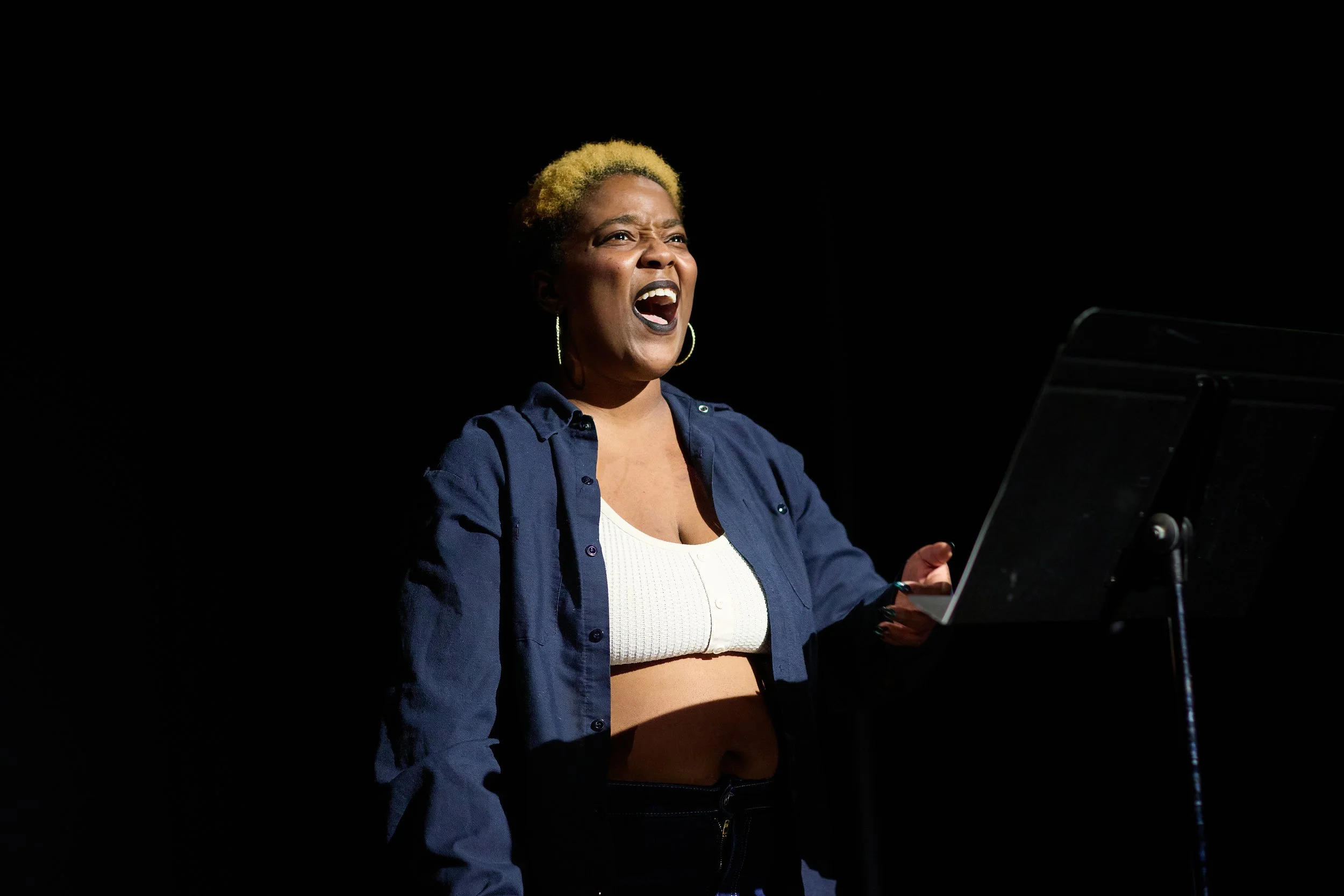 Une femme chantant ou parlant passionnément devant un microphone sur scène, vêtue d'une chemise bleue, de vêtements blancs et portant des boucles d'oreilles.
