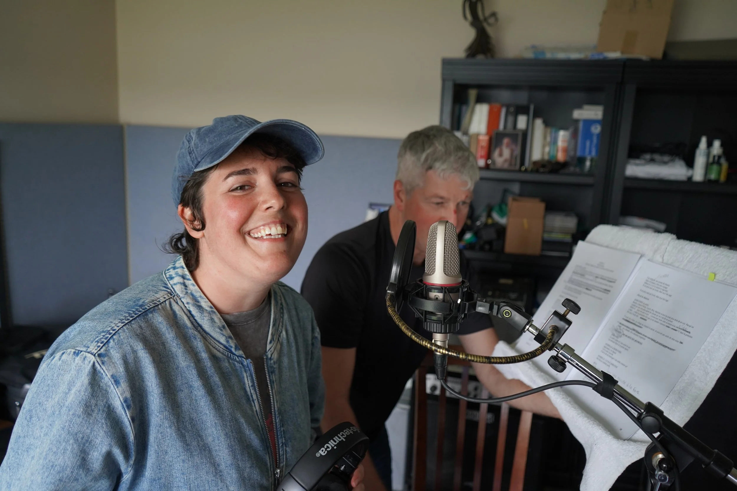 Deux hommes dans un studio d'enregistrement, un avec un chapeau en jean souriant et l'autre concentré, utilisant un microphone sur un support