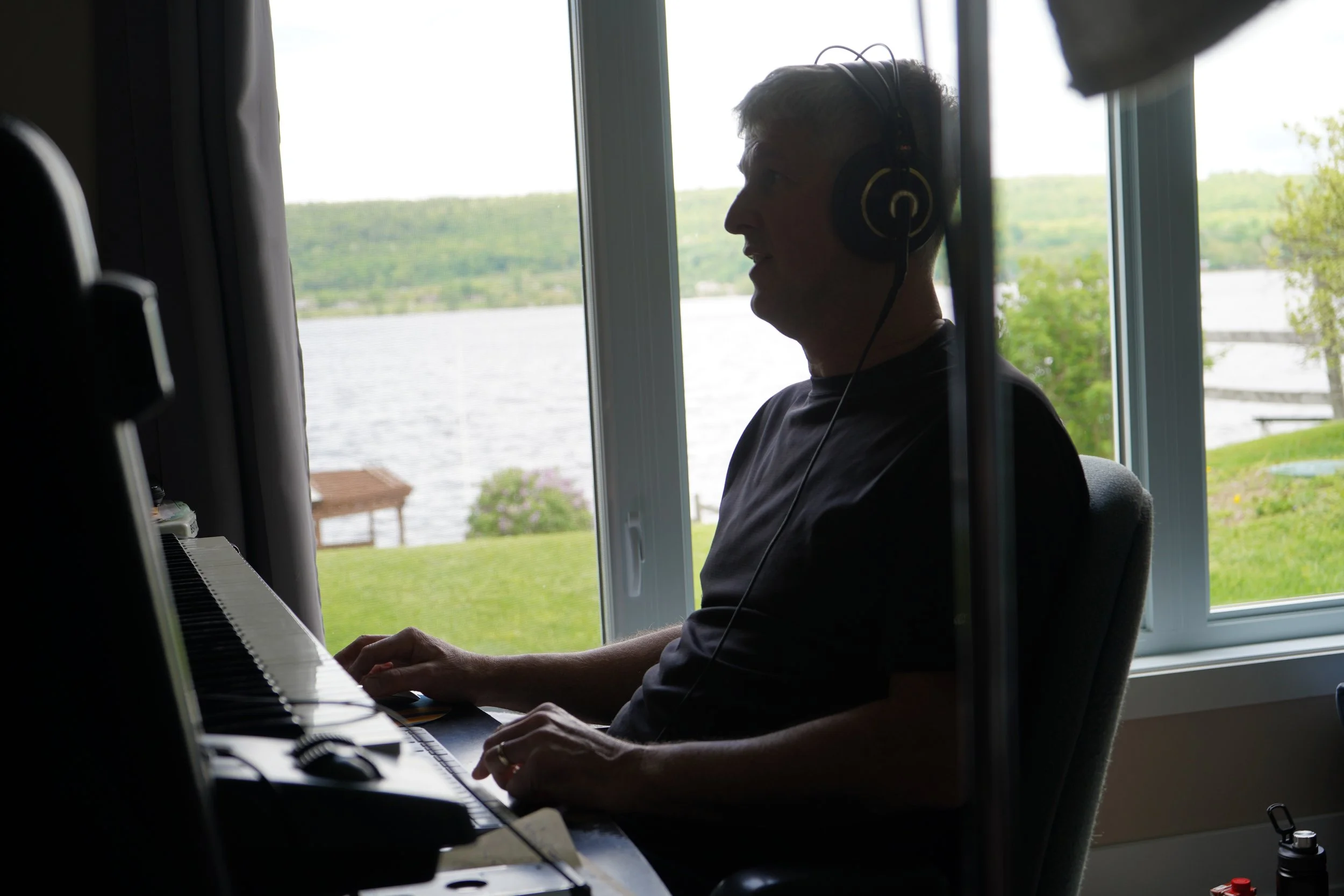 Un homme jouant du piano dans une pièce près d'une fenêtre. Il porte un casque et regarde par la fenêtre vers un paysage verdoyant avec un lac ou une rivière en arrière-plan.