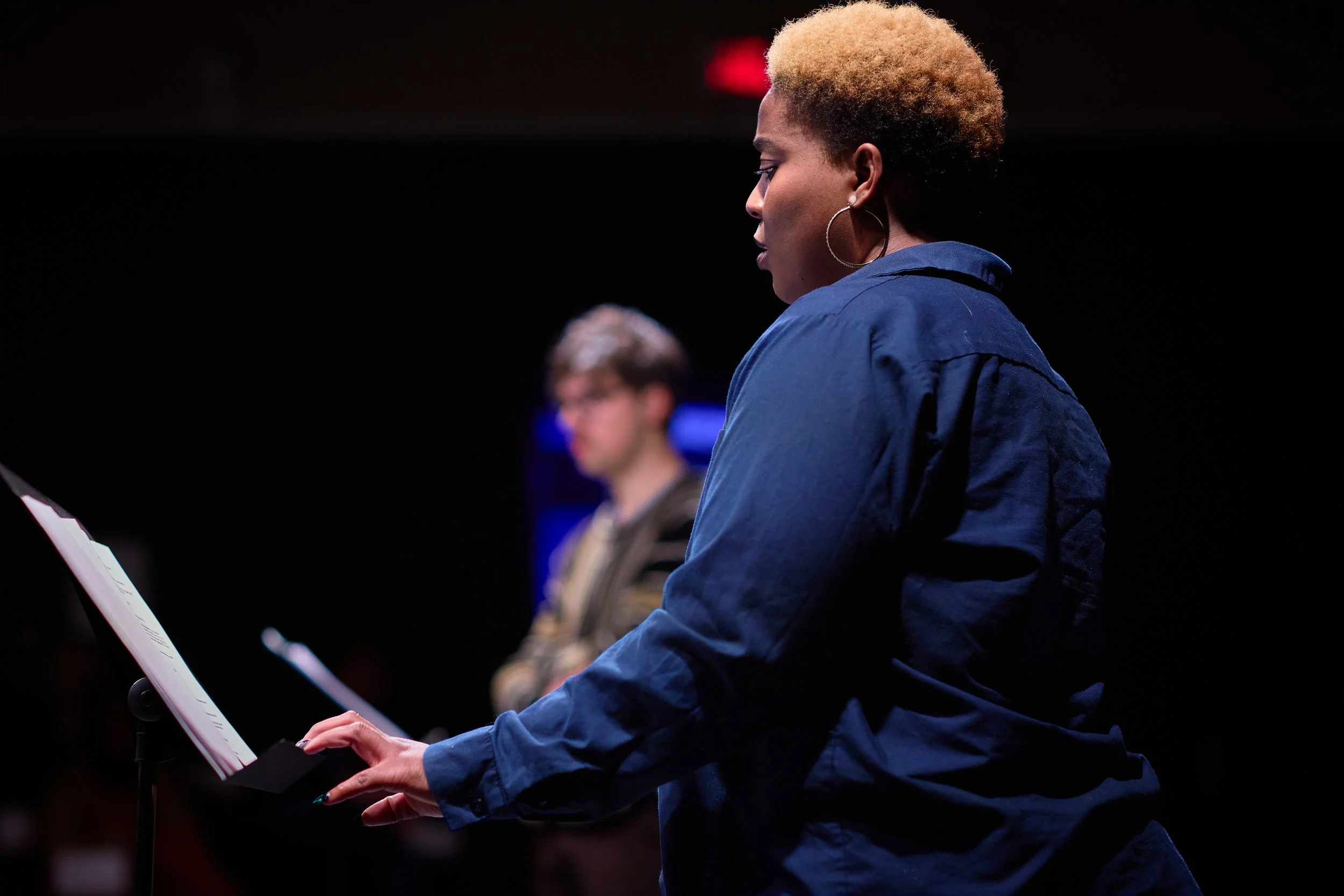 Une femme blonde lisant une partition lors d'une répétition musicale, avec un homme flou en arrière-plan dans une scène sombre.