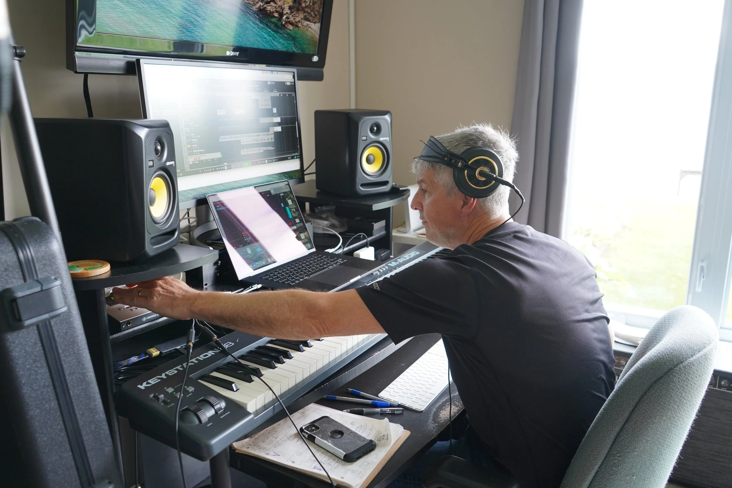 Un homme avec des cheveux gris portant un casque d'écoute travaille dans un studio de musique avec des haut-parleurs, un clavier numérique, un ordinateur portable et un grand écran.
