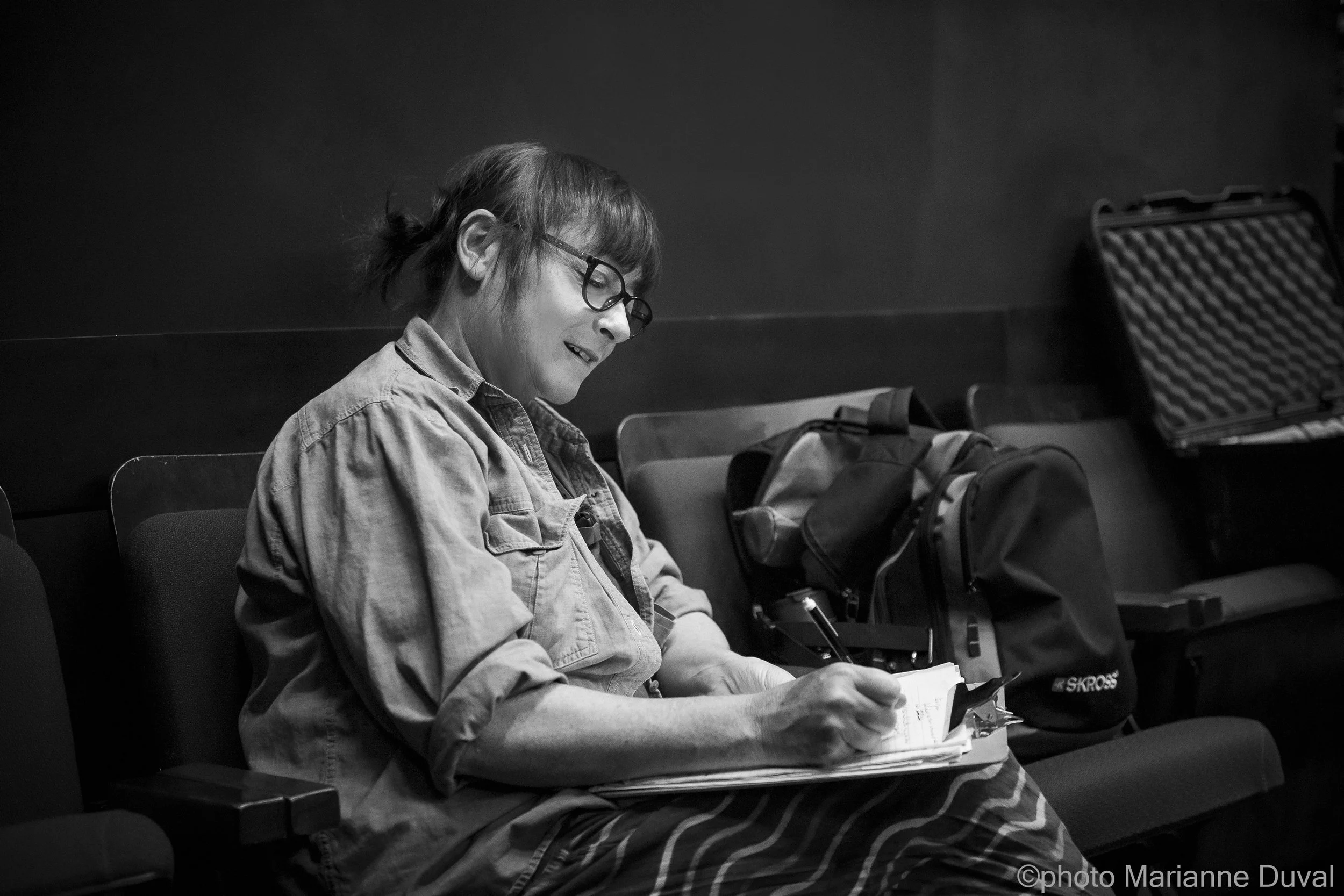 Femme assise portant des lunettes, écrivant dans un carnet, avec des sacs à dos à côté, en noir et blanc.