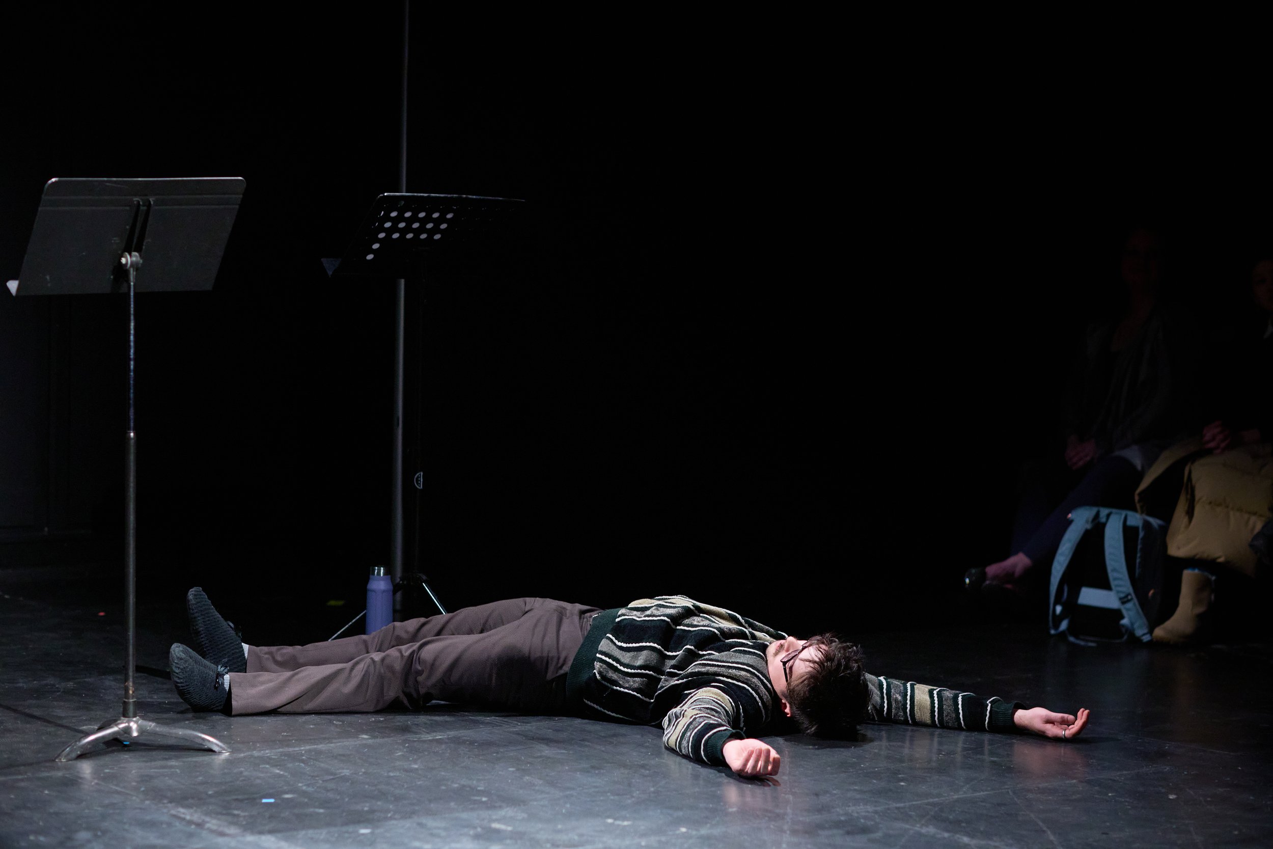 Une personne allongée sur le sol d'une scène théâtrale, apparemment en train de jouer ou de faire une performance, avec deux autres personnes assises en arrière-plan, dans une pièce sombre et un peu mystérieuse.