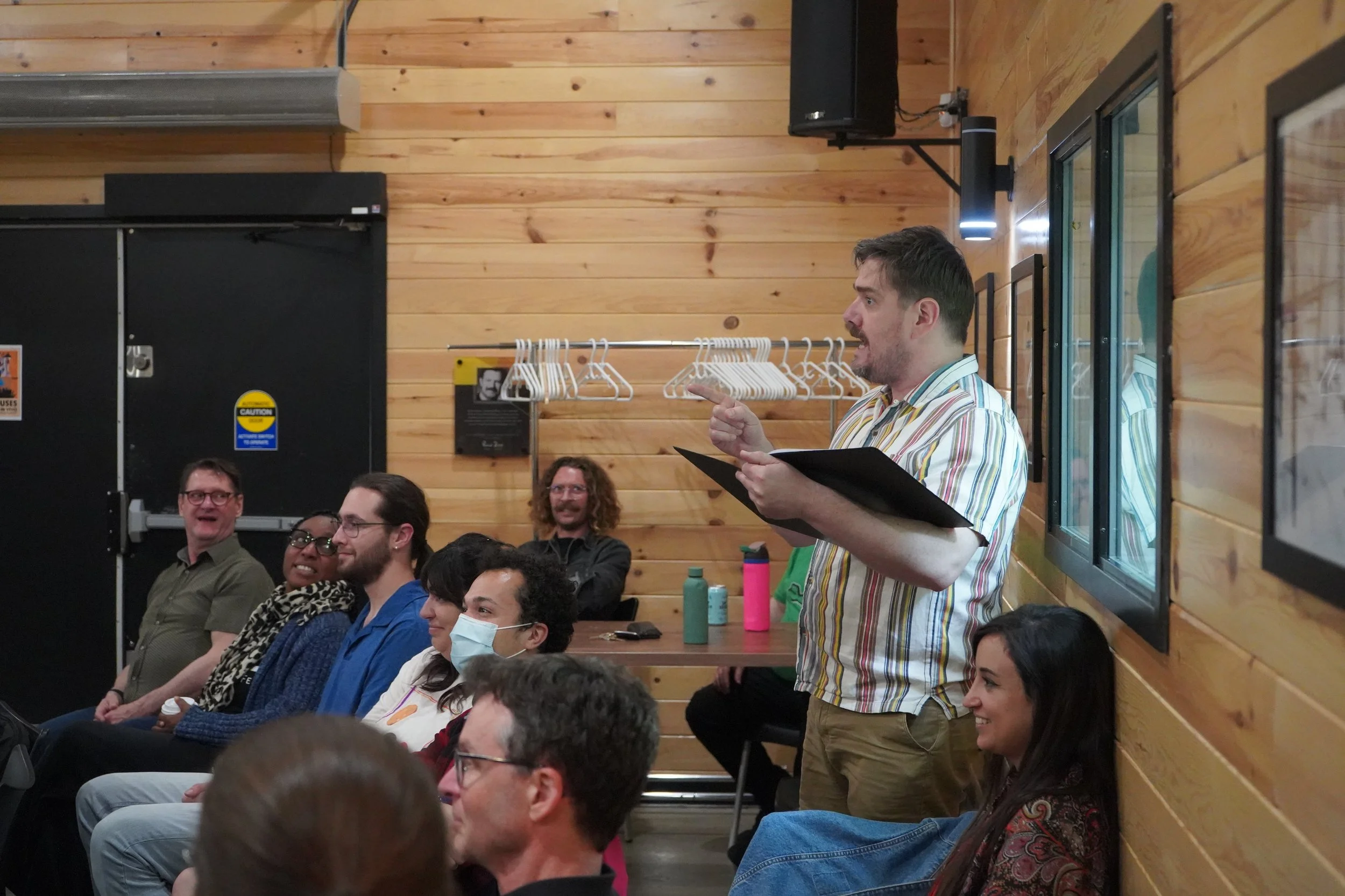 Un homme parlant à un groupe de personnes assises lors d'une réunion ou discussion dans une salle en bois, diverses personnes écoutent attentivement, certaines portent des masques et des vêtements décontractés.