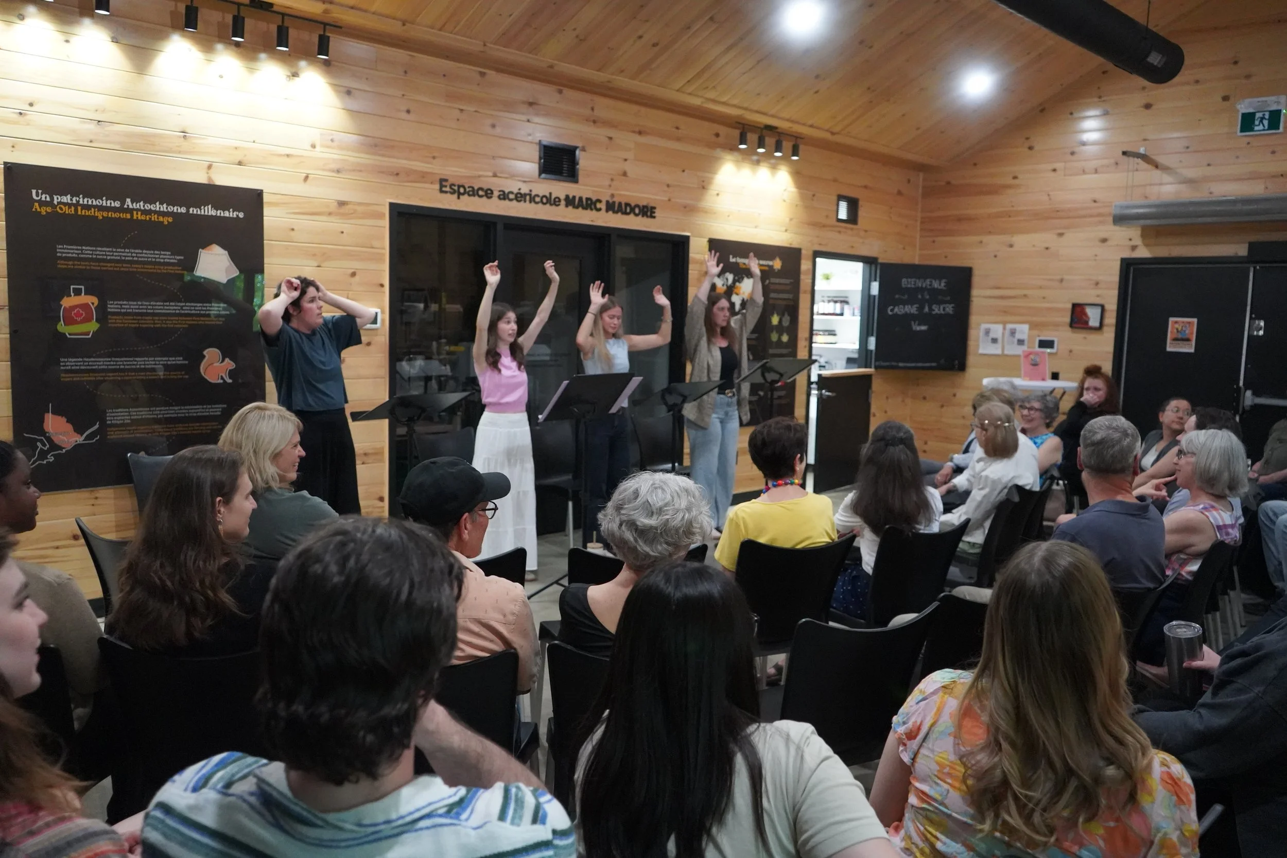 Présentations ou conférence dans une salle en bois avec un groupe de personnes assises et quatre femmes au centre qui semblent parler ou faire une démonstration.
