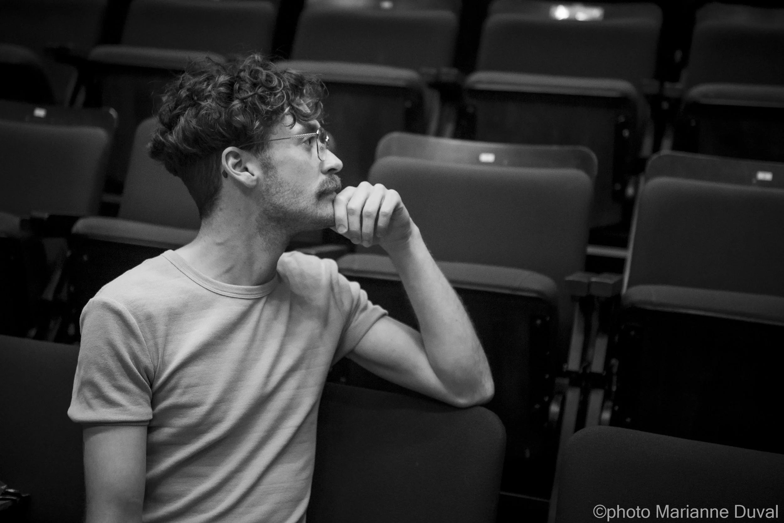 Un homme aux cheveux bouclés et portant des lunettes, assis dans une salle vide, semble réfléchir avec sa main sous le menton.