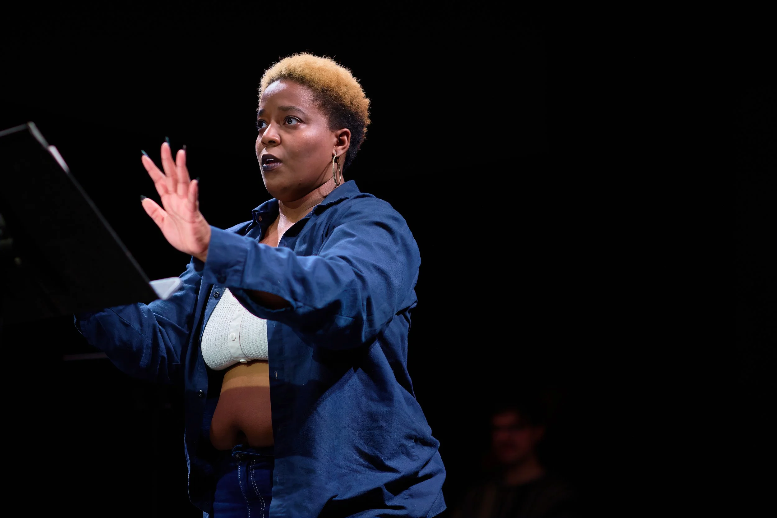 Une femme de couleur avec des cheveux courts blond foncé, portant une veste bleue et une chemise blanche, semble donner un récital ou faire une intervention avec des mains levées. Elle est sur scène contre un fond noir.