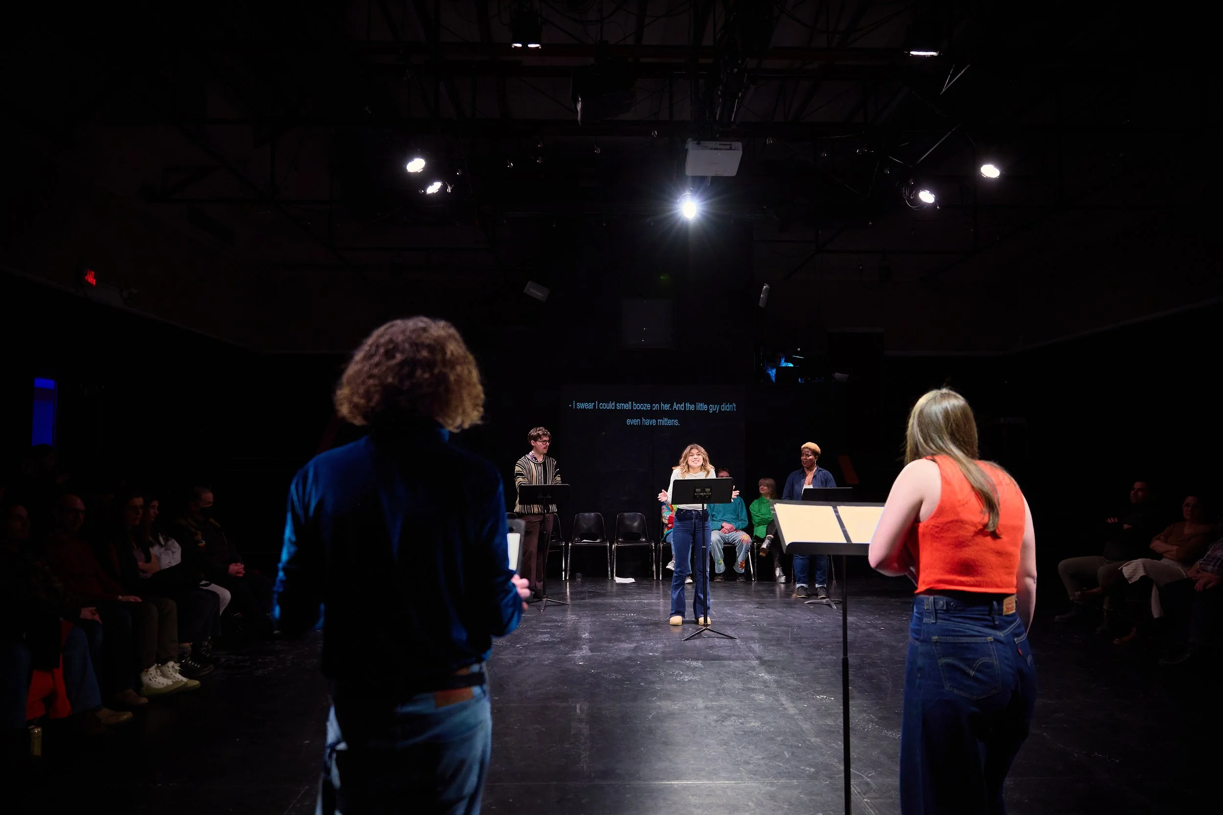 Une scène de théâtre ou de répétition avec plusieurs personnes, dont une femme au centre parlant ou chantant, entourée de ses compagnons, avec un public en arrière-plan et une scène sombre.