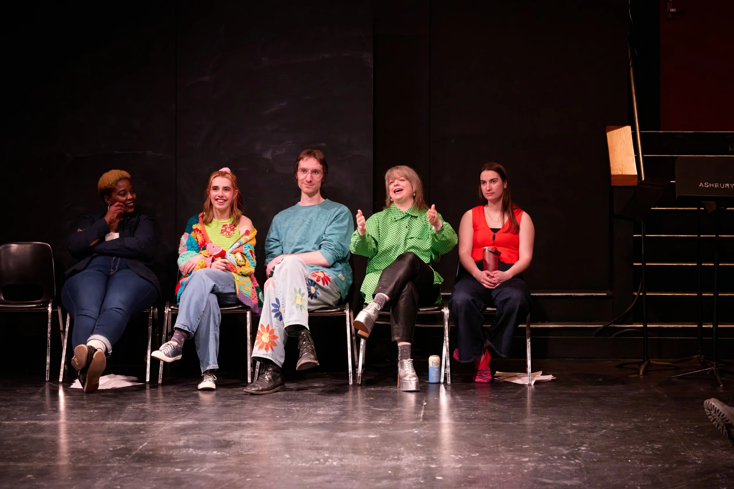 Cinq jeunes assis en ligne sur scène, discutant ou en pause, avec un piano à queue noir à droite et un mur noir en arrière-plan.