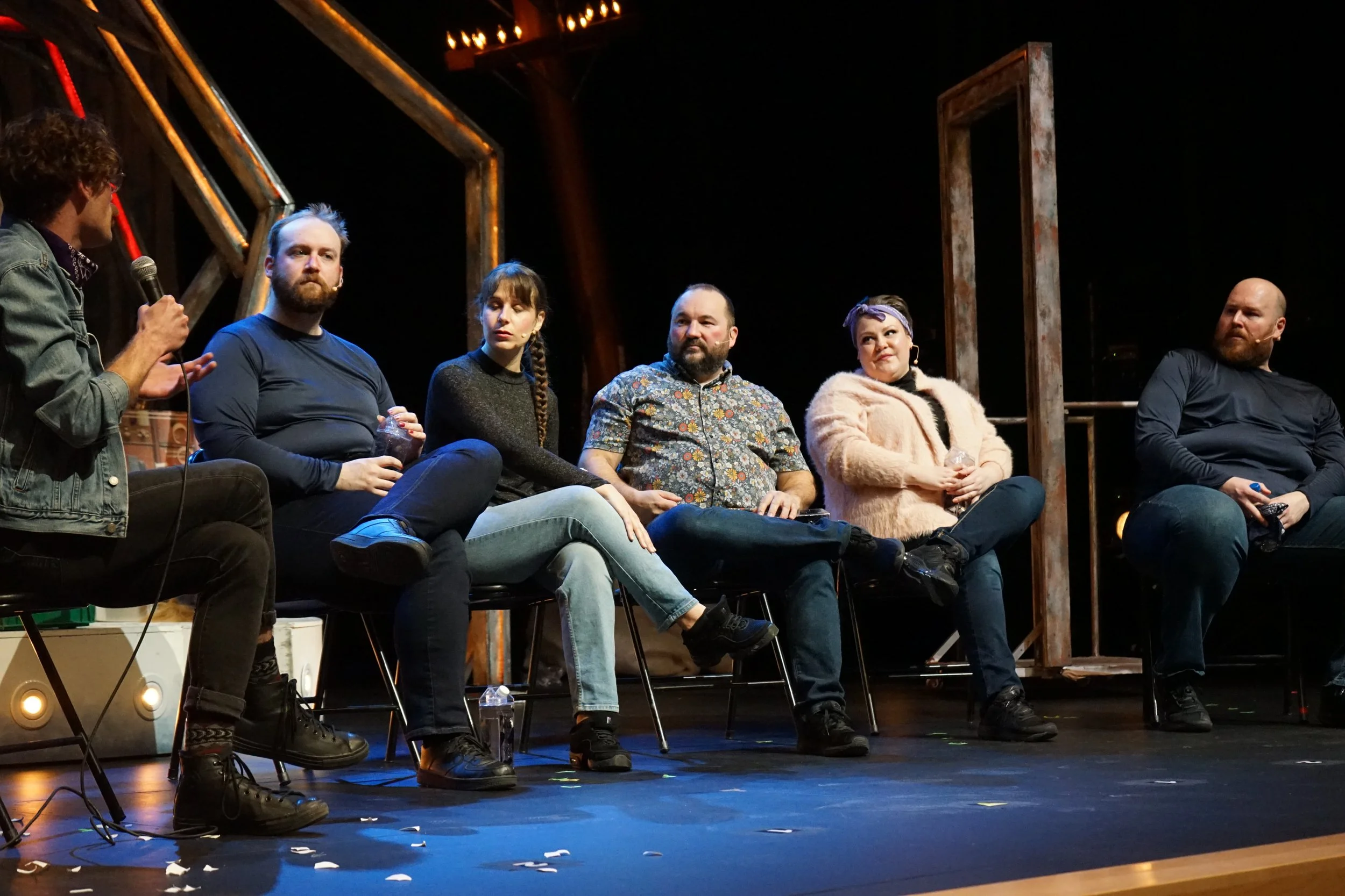 Groupe de six personnes assises sur scène lors d'une discussion, avec un intervenant tenant un micro, décor industriel avec des éclairages chauds.