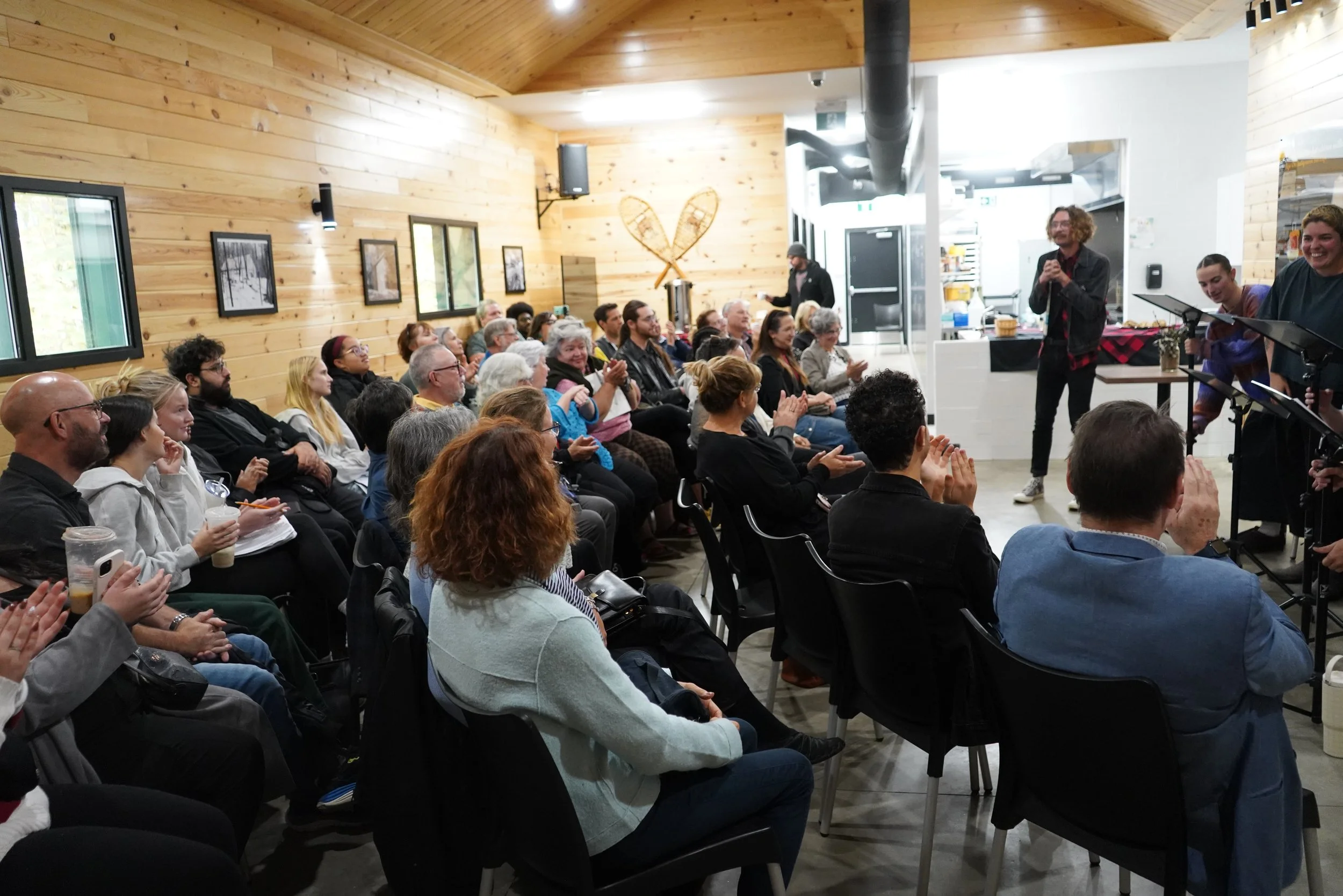 Une salle avec des personnes assises regardant un concert ou une présentation, avec un mur en bois et une décoration de patins à glace en bois au fond, et un groupe de musiciens ou de conférenciers devant eux.
