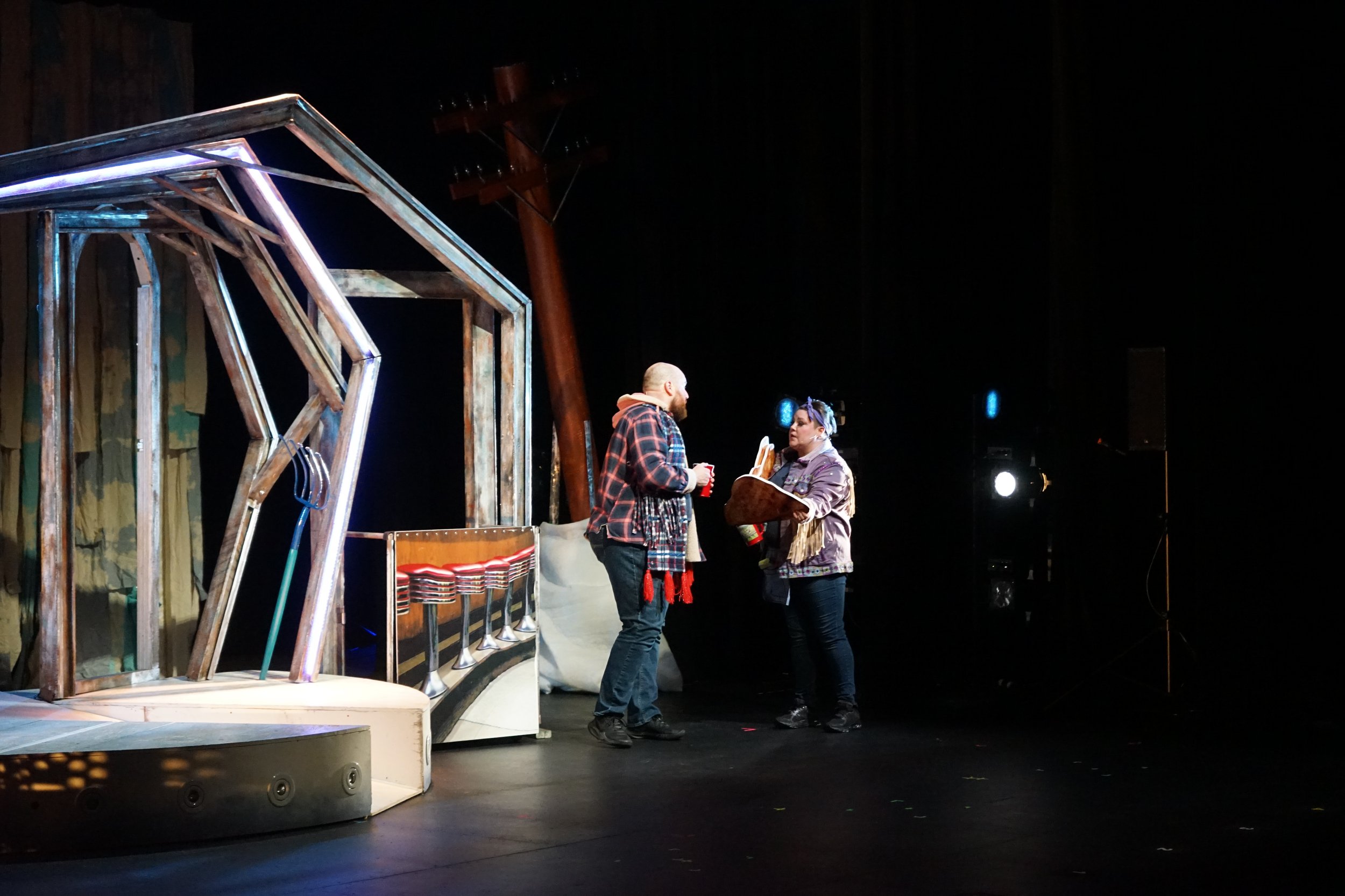 Deux personnes discutent sur une scène de théâtre, avec un décor en bois.