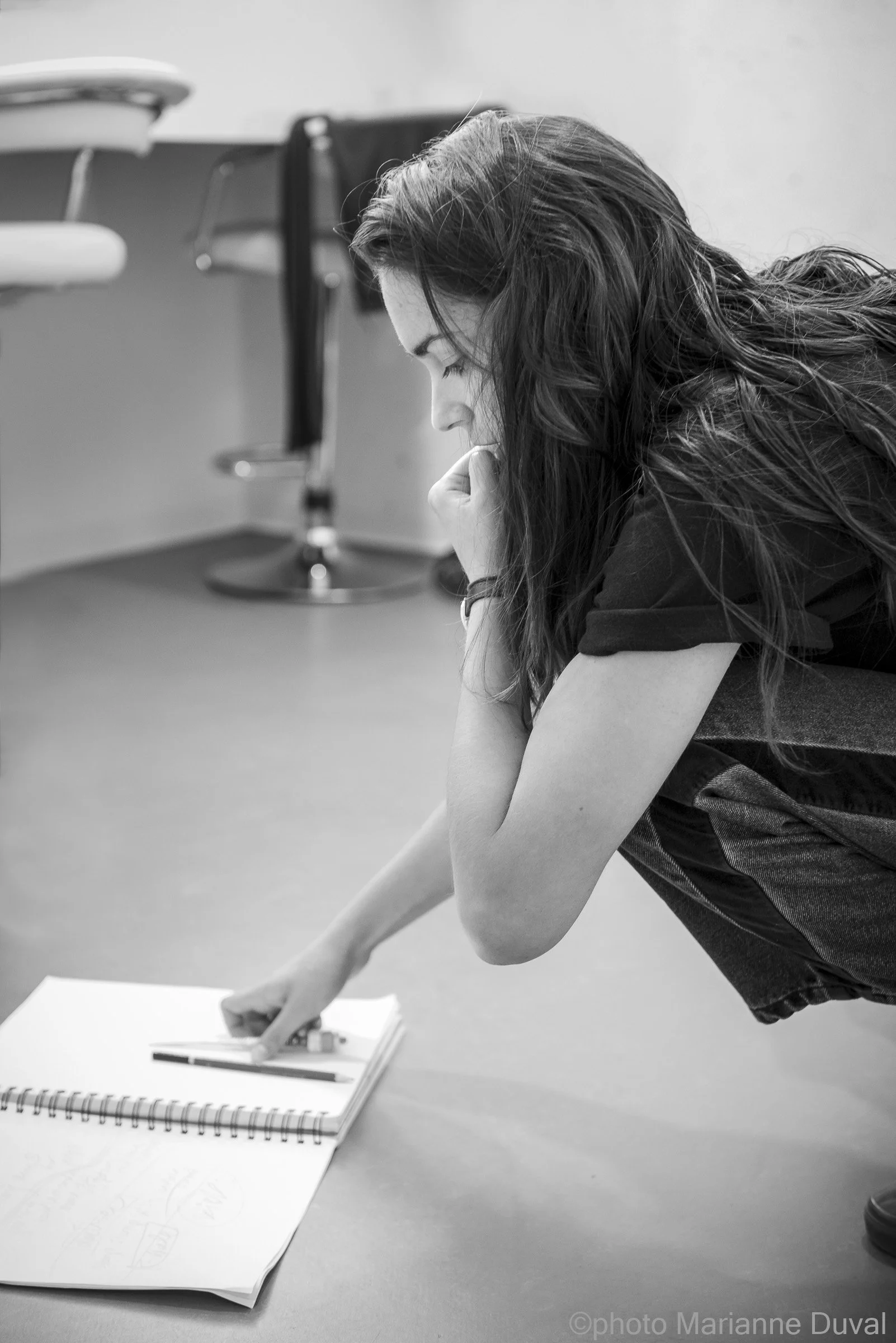 Jeune femme concentrée, pliée en position de squat, pointant du doigt un cahier ouvert avec des crayons, dans une pièce.