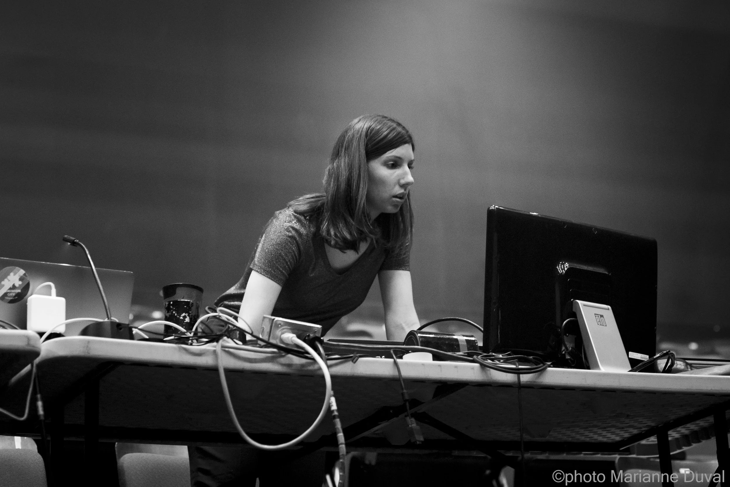 Une femme regarde un écran d'ordinateur sur une table, entourée de câbles et d'équipements technologiques, dans un environnement sombre.