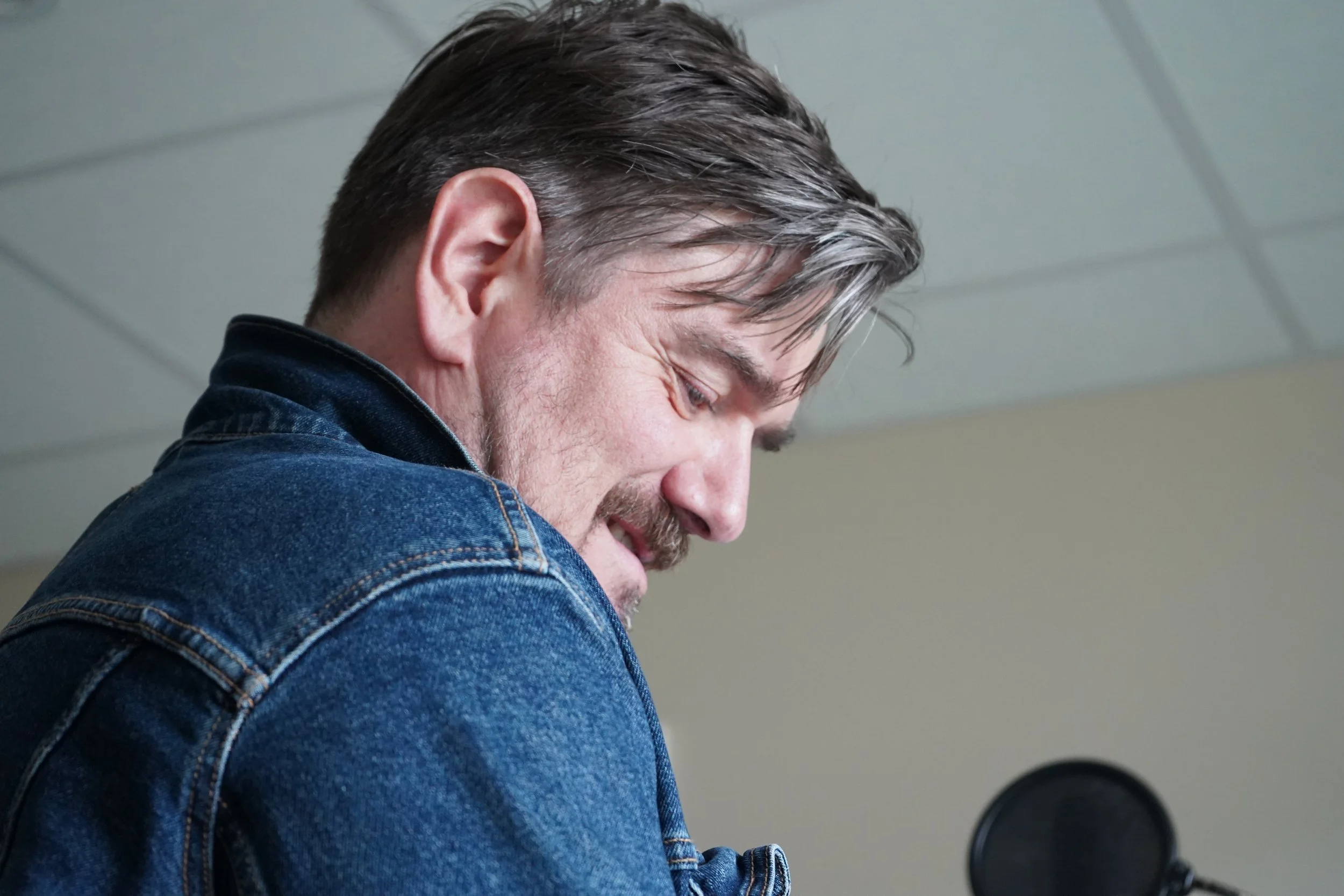 Un homme avec des cheveux foncés et une barbe porte un jean bleu clair et regarde vers le bas