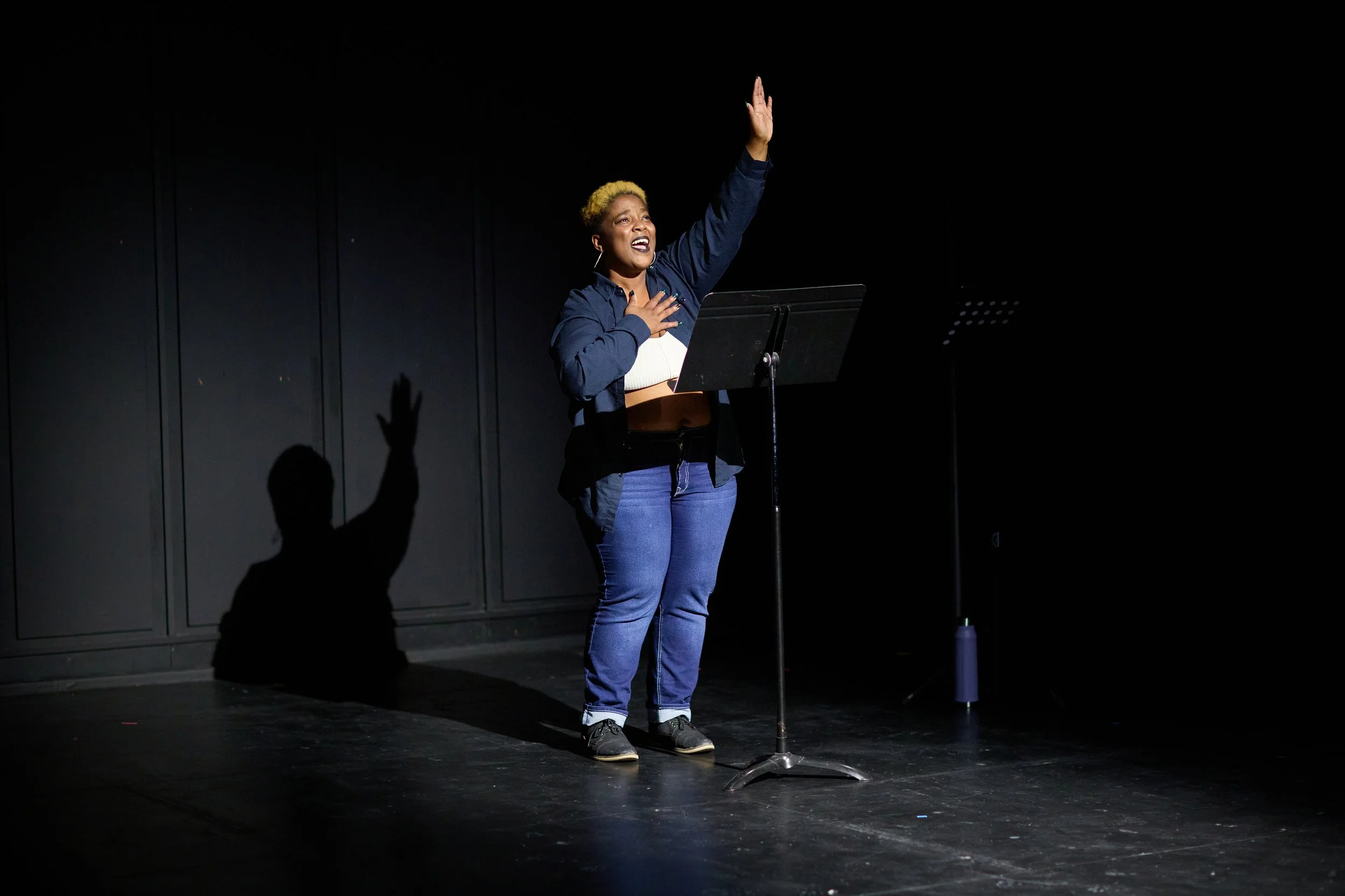 Une femme sur scène avec un micro, l'air expressive et joyeuse, en train de chanter ou de parler, avec un light qui met en valeur sa présence.